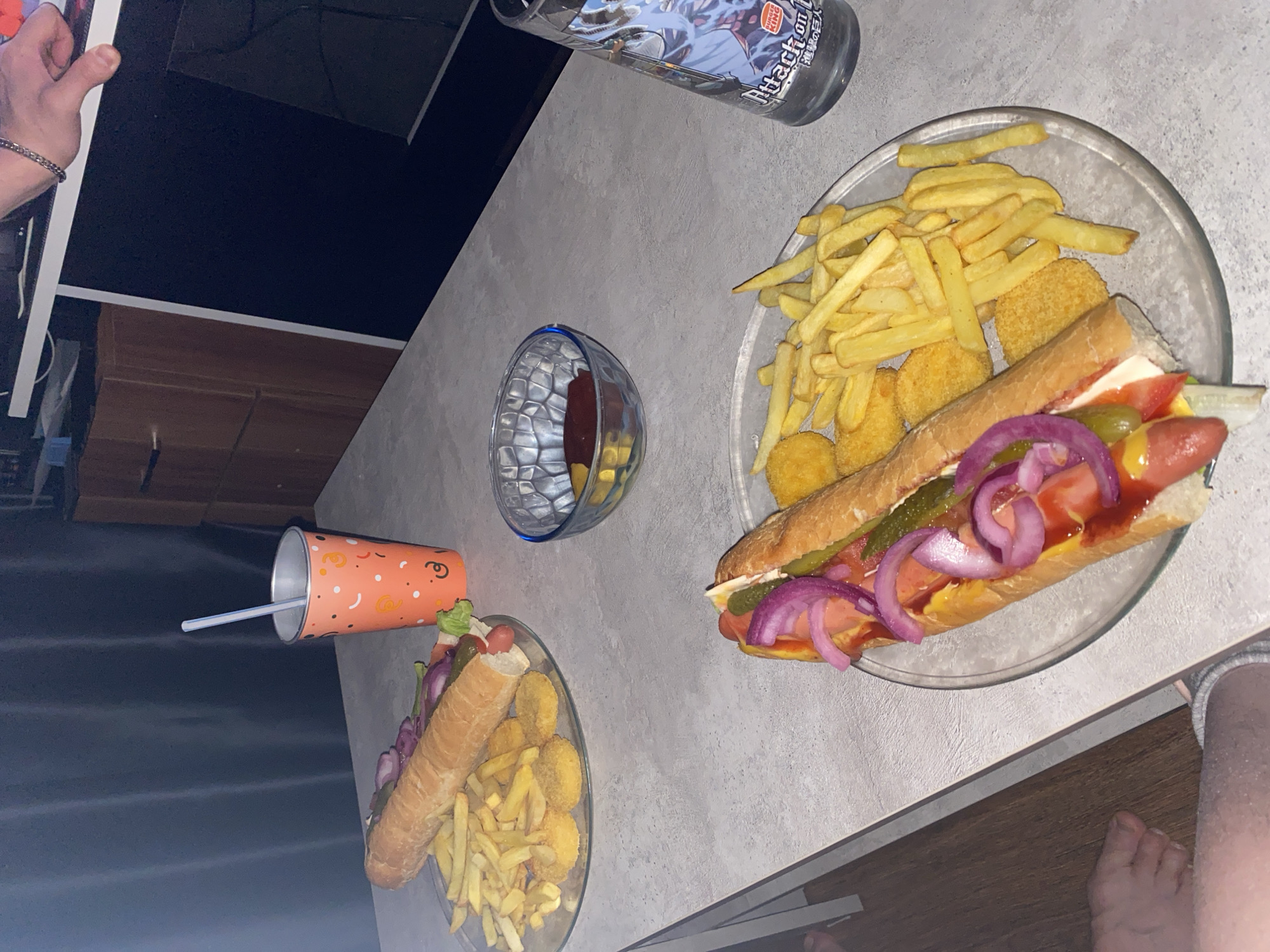 A cluttered tabletop displays two plates each holding a long hot dog, french fries, and chicken nuggets. The hot dogs are topped with red onions, pickles, ketchup, and mustard. A clear drinking glass containing a red liquid sits between the two plates. Above the plates, there is a water bottle and below the plates, there is an orange cup with a straw. Human limbs are seen at the edges of the image, indicating that people are present at the table. The environment suggests an indoor setting. There is no information to place this image in Izhevsk, Russia.