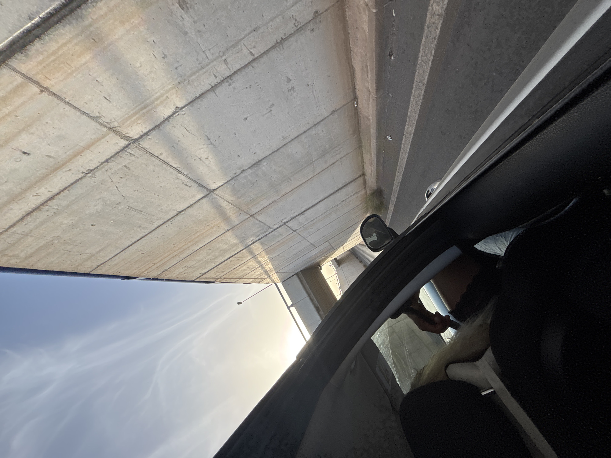 The image depicts a view from inside a car, looking out towards a concrete overpass and the sky. The car's interior is visible in the foreground, with the dark seats and a portion of the dashboard. A person's hand, with blond hair visible, is reaching towards the steering wheel. Outside the car, a massive concrete structure, likely part of a highway or bridge, dominates the right side of the frame. This structure appears to be segmented and shows signs of wear and staining. The sky is blue with wispy white clouds, and the sun is visible, casting a bright glow. Below the overpass, a road is visible with lane markings. The overall scene suggests the car is traveling on or under a large infrastructure project. The location is identified as Orihuela, Spain.