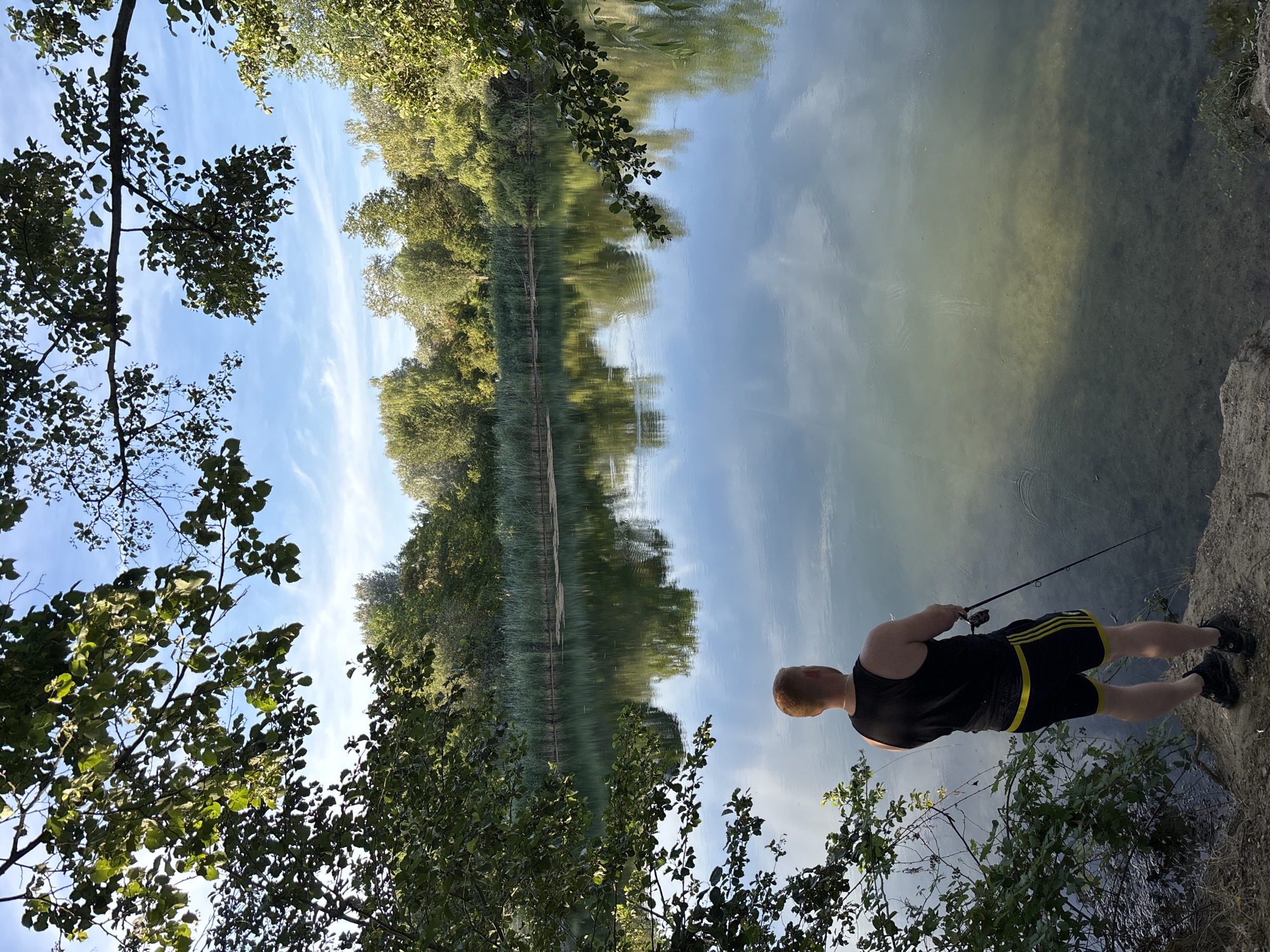 The image shows a man fishing on the shore of a lake or pond. He is standing on a dirt embankment, facing the water. He is wearing a black tank top and black shorts with yellow stripes. He is holding a fishing rod. The water is calm and reflects the trees and sky. In the background, there is a line of reeds and more trees. The sky is blue with some white clouds. The image appears to have been taken on a sunny day. The location is likely the Baggersee in Ludwigshafen am Rhein, Germany, a popular recreational area.