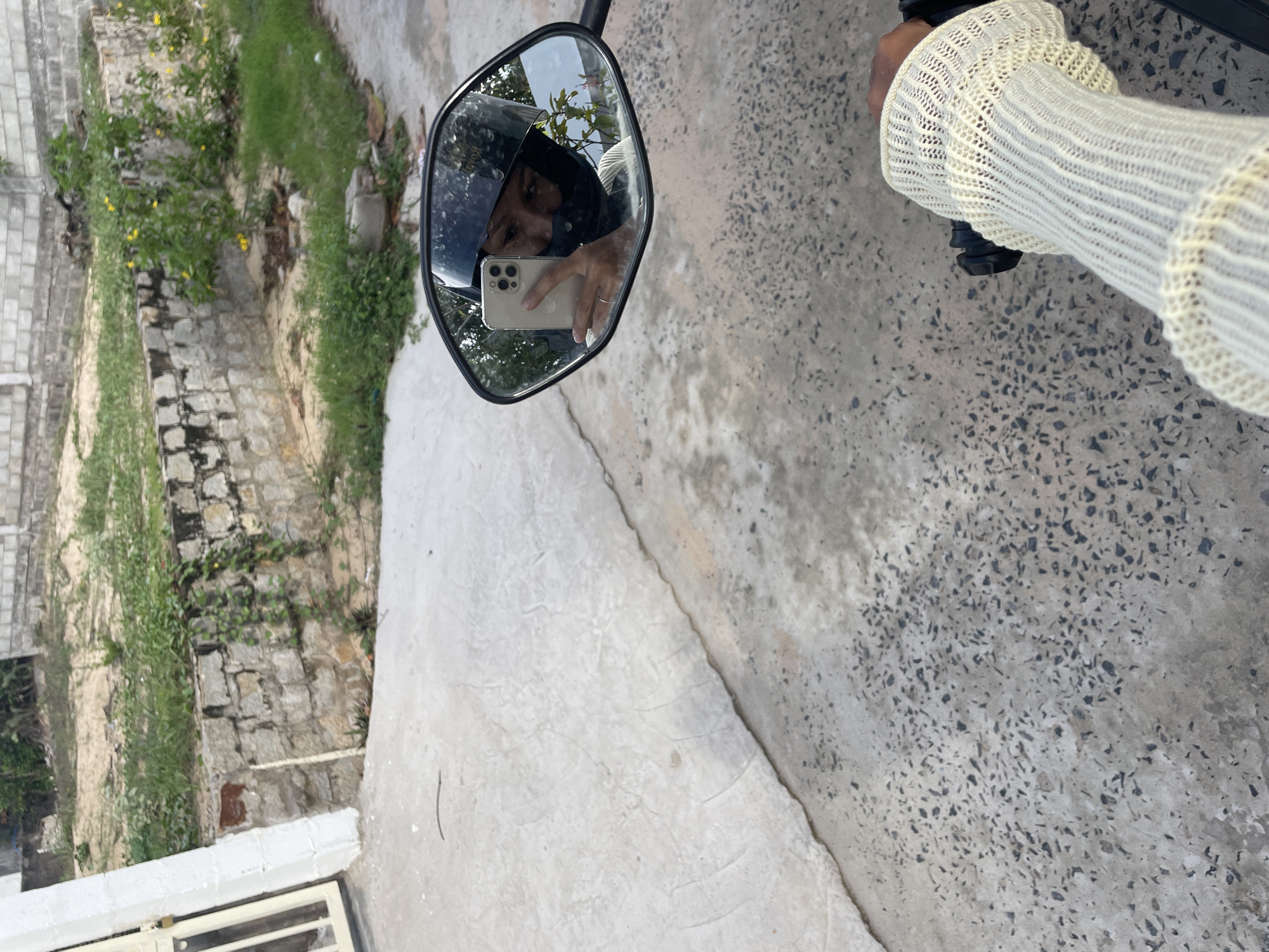 The image captures a unique perspective of a person taking a selfie, reflected in a motorcycle's rearview mirror. The mirror is prominently featured in the central-left area, showing a person wearing a black helmet and holding an iPhone with multiple cameras to capture their reflection. The person's face is partially visible behind the phone. Faint text, possibly "HONDA," is discernible on the helmet in the reflection.
To the right of the mirror, the person's arm is visible, clad in a cream or light-yellow long-sleeved top with a distinct knitted pattern, holding onto a motorcycle handlebar. The arm suggests a person with darker skin tone.
The scene is set outdoors during daylight hours, indicated by the bright reflections and clear visibility. The ground surface features a light grey concrete mixed with dark aggregate, transitioning to smoother concrete areas. The background, particularly to the left, reveals a lush environment with green vegetation, including bushes dotted with small yellow flowers, and a weathered stone wall structure. Parts of a white fence or gate are visible in the bottom-left corner. The overall impression is a casual outdoor moment in what appears to be Phu Thuy Ward, Vietnam, under sunny conditions.