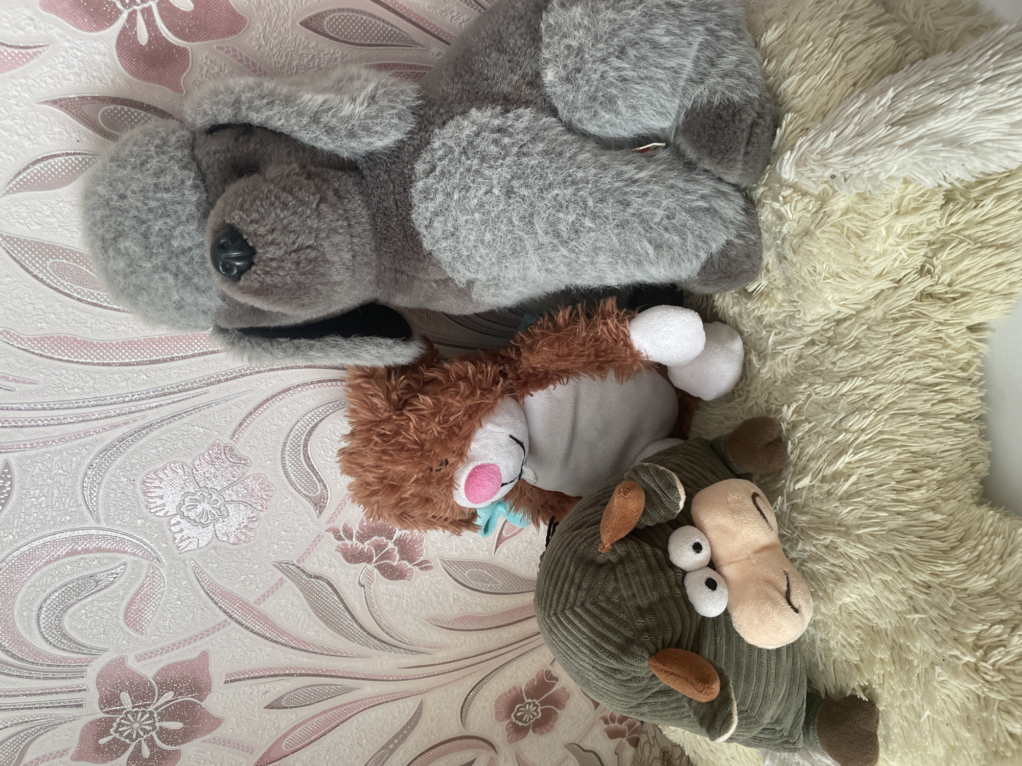 Three stuffed animals are displayed on a fluffy cream-colored rug in front of a floral wallpapered wall. On the left is a green, corduroy bull with large, expressive eyes. In the center is a brown, shaggy-haired creature with a white belly and a pink nose. To the right is a large, grey poodle with a fluffy coat and floppy ears. The poodle is the tallest of the three and sits upright. The bull is slumped over, looking towards the viewer. The brown creature is leaning slightly to the right. The background wallpaper features a repeating pattern of stylized pink and grey flowers and leaves with subtle glitter accents. The lighting appears to be from a soft, diffused source, casting minimal shadows.