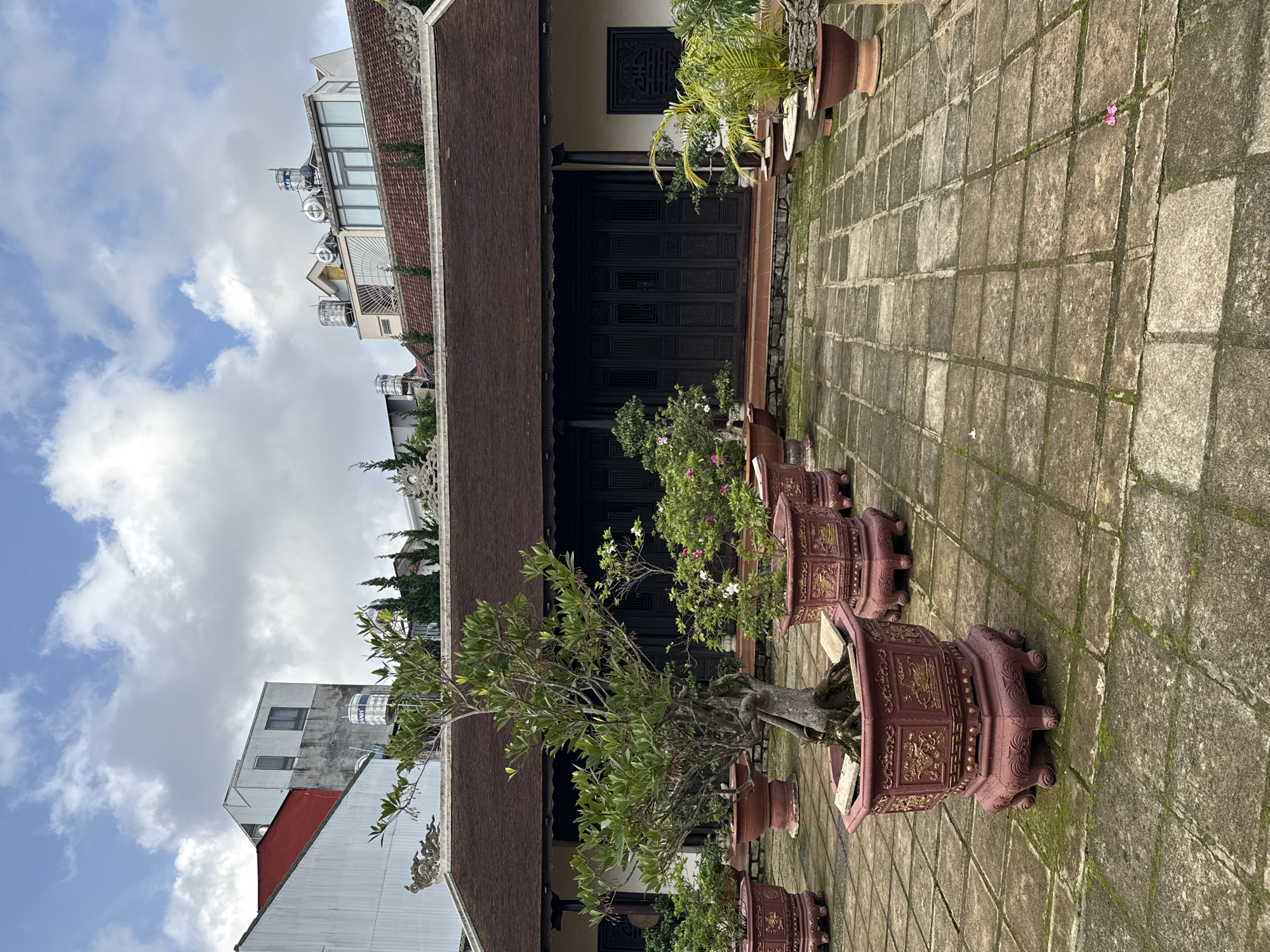This image, set in Da Lat, Vietnam, presents an outdoor courtyard scene featuring traditional architecture contrasted with modern buildings under a partly cloudy sky.
In the foreground, a courtyard is paved with large, weathered square and rectangular stone tiles, many showing signs of moss growth in their seams. Lining one side of this courtyard are several substantial, ornate reddish-brown planters. These elaborate pots feature intricate gold patterns and decorative feet, housing a variety of plants. A prominent planter holds a mature, gnarled tree with green leaves, reminiscent of a large bonsai. Other pots contain smaller bushes with white and pink flowers, and some palm-like foliage.
The main building bordering the courtyard exhibits traditional Vietnamese architectural elements. It features a dark, multi-layered tiled roof with decorative carved eaves along its edge. Beneath the roof, a light-colored wall frames a large set of dark, intricately carved wooden double doors. Above these doors, a smaller square wooden panel with a lattice-like design is embedded in the wall.
Beyond the traditional building, in the background, several modern multi-story structures are visible. These buildings have plainer facades with windows and feature numerous silver water tanks on their rooftops. Visible text on some of these tanks includes "TIEN AN" and "SAO VANG".
The sky above is bright blue with scattered white and grey clouds, indicating daytime. No people or specific activities are depicted, giving the scene a serene and static quality. The image captures a blend of old and new architectural styles typical of a developing urban landscape.