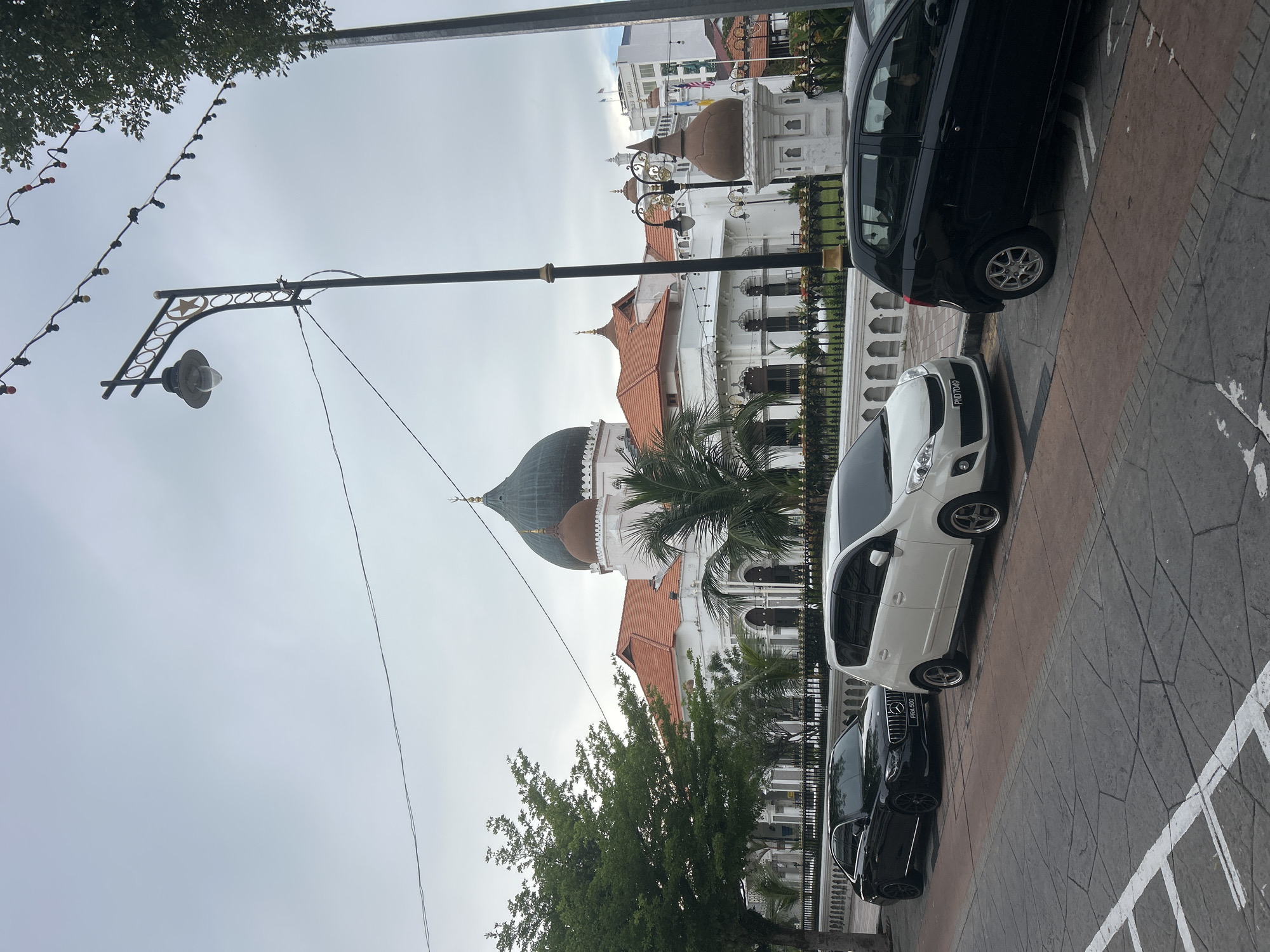 The image shows the Kapitan Keling Mosque in George Town, Malaysia, on a cloudy day. In the foreground, three cars are parked along a street with patterned paving stones. From left to right, they are a black sedan, a white hatchback, and a black hatchback. The mosque, a prominent white building with a large green dome and several smaller brown domes, is visible behind a decorative white fence and lush green trees. A streetlamp with a star design stands to the left of the frame, and a string of colorful lights hangs overhead. The sky is overcast, contributing to a diffused light.