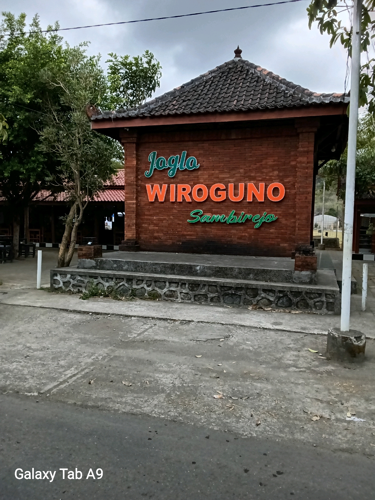 An outdoor shot shows the entrance to a place called "Joglo WIROGUNO Sambirejo" in Kelutan, Indonesia. The entrance is made of brick and has a tiled roof. The name of the place is displayed in large orange and green letters. There are trees and foliage around the entrance, and a paved area in front of it. The sky is cloudy. The image also has a watermark that says "Galaxy Tab A9".