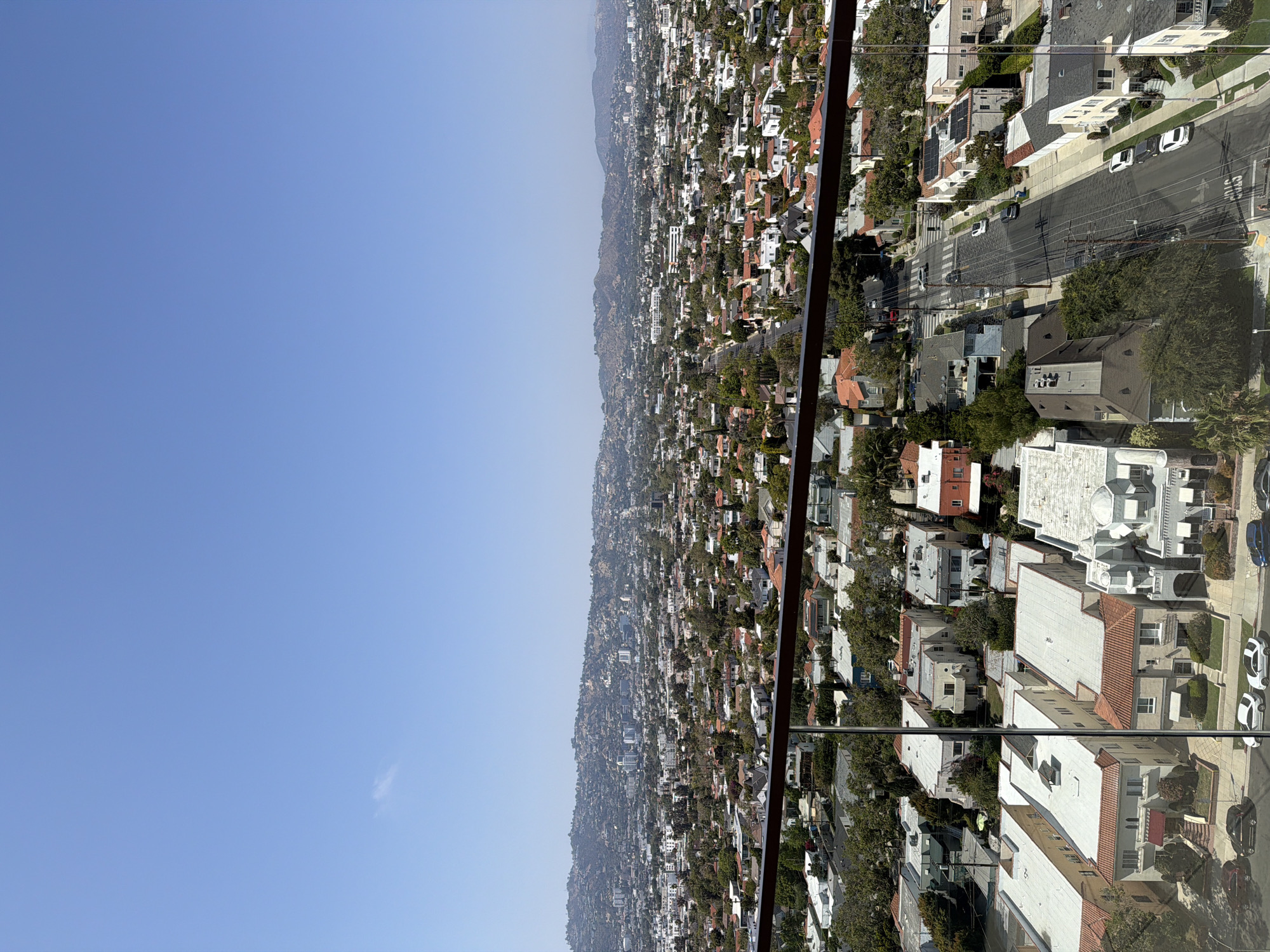 The image shows a view of a densely populated residential area in the hills of Los Angeles, California, United States of America. The scene consists of numerous houses with varying architectural styles and roof colors, interspersed with trees and vegetation. Streets with parked and moving cars can be seen running through the residential area. The sky is clear blue. The view is taken from an elevated position, possibly from a window, and includes a metal railing in the foreground.