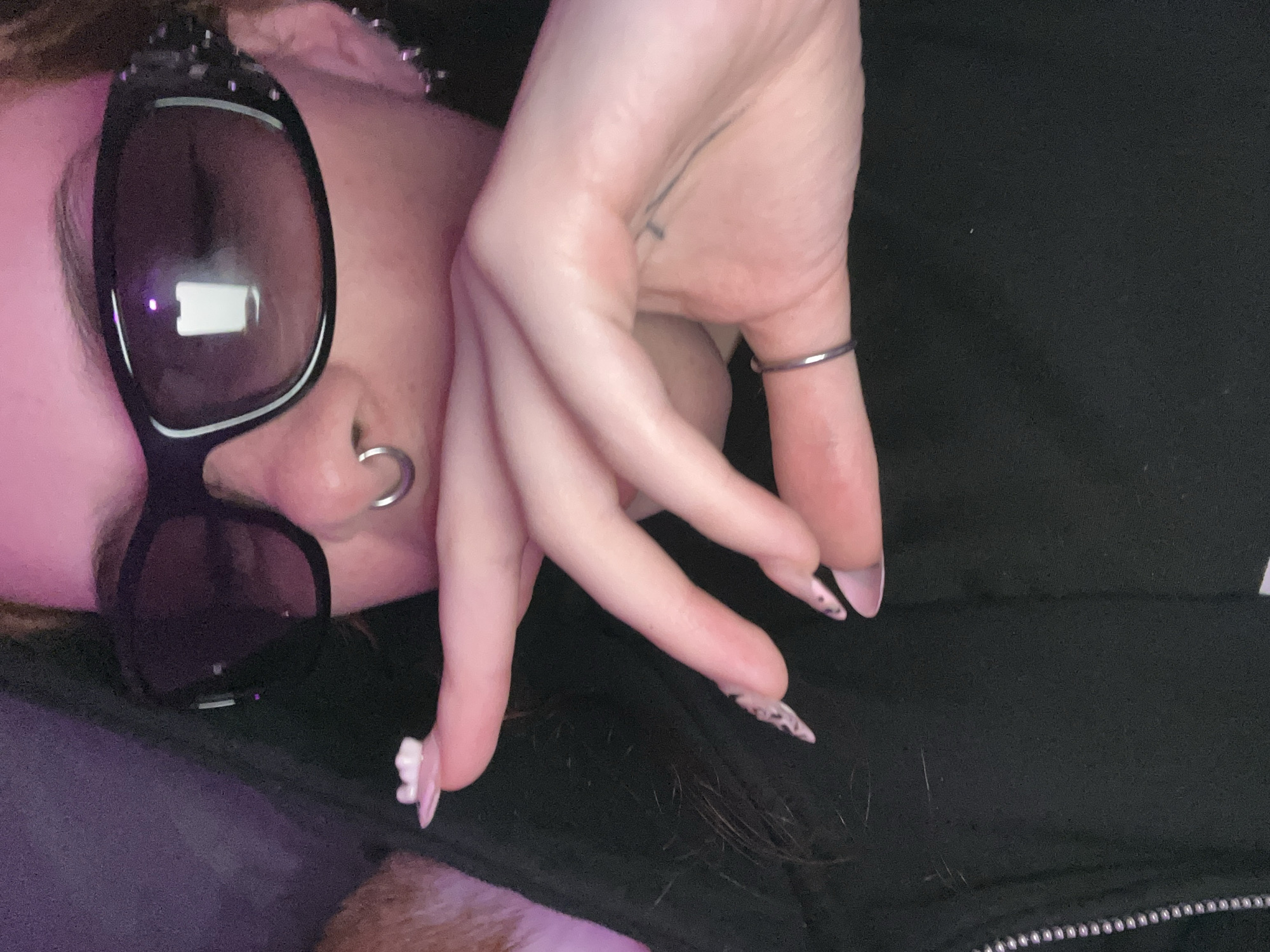 A selfie-style photograph shows a person wearing sunglasses and a nose ring. The person's left hand is raised near their face. The nails on the fingers are decorated with a pink and white design. The background is a black fabric, possibly clothing. A portion of a zipper can be seen. The location is not discernible from the image. There is no indication that the image was taken in Buenos Aires, Argentina.