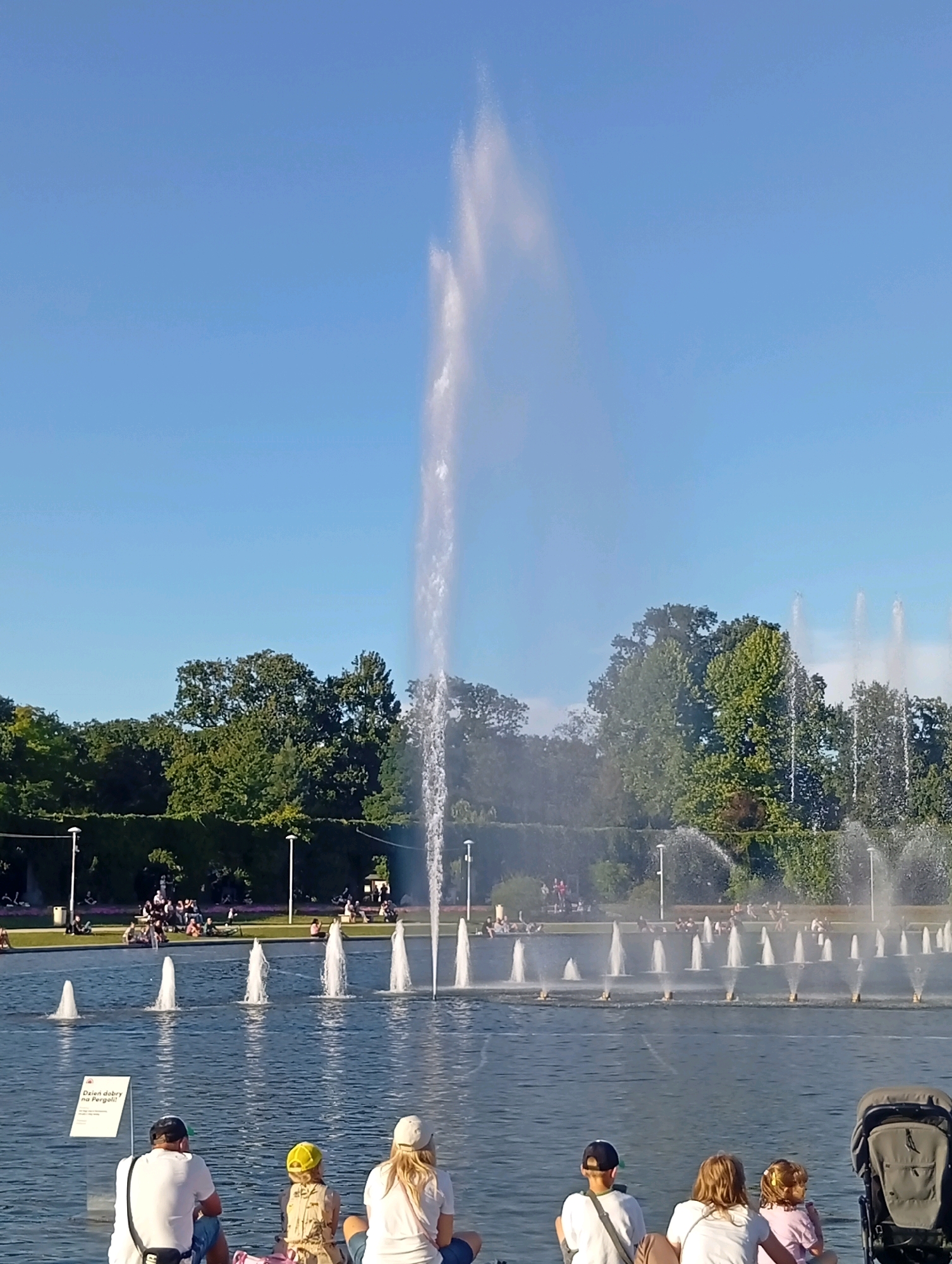 This image captures a scenic view of the multimedia fountain in Wrocław, Poland, on a clear day. In the foreground, four individuals are seated on the edge of the water. A man in a white t-shirt and black baseball cap faces away from the camera, a woman in a white t-shirt and white baseball cap is to his right, and a child in a yellow hat is between them. To the right of the woman is another individual, also facing away, and a child to their right. A baby stroller is positioned to the far right. A white sign with Polish text is visible in the lower left corner.
The middle ground features a large body of water with numerous active fountains, creating a dramatic spray against the backdrop of lush green trees and vegetation. In the background, more people are scattered across grassy areas and walkways, enjoying the park. The sky is a vibrant, cloudless blue. The overall atmosphere suggests a pleasant, leisurely day at a popular public attraction.