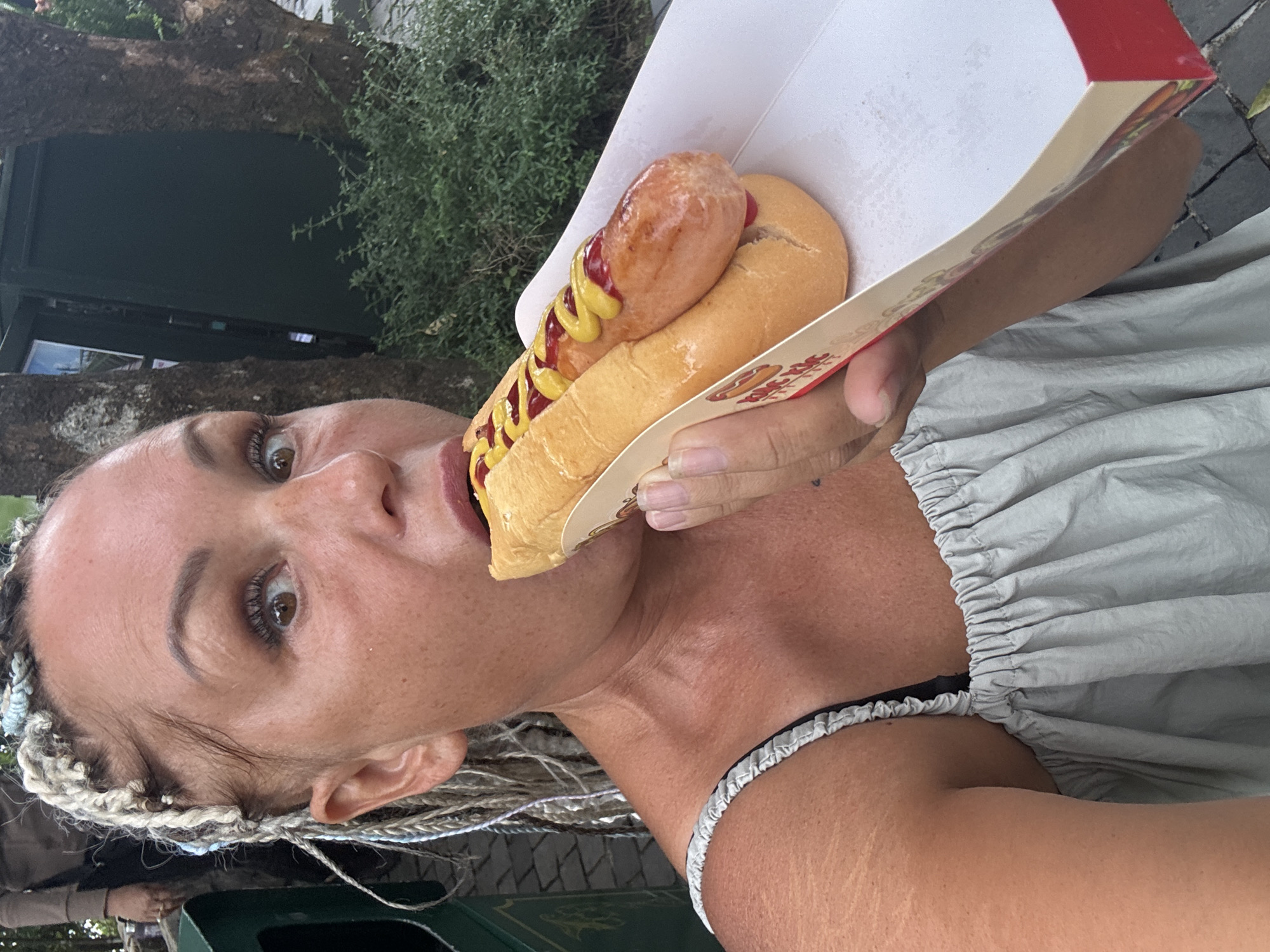 A woman with tan skin and light brown eyes is holding a hot dog in a white and red cardboard holder up to her mouth. The hot dog has a long, oval-shaped sausage, mustard, and ketchup on a bun. The woman has blonde braids and is wearing a light gray shirt. She has a silver bracelet on her left wrist. The background is blurry, but it appears to be an outdoor setting with a tree and some green foliage. There is also a dark green structure or object visible in the bottom left corner. There is no evidence that this photo was taken in the city of Đà Nẵng, Vietnam.