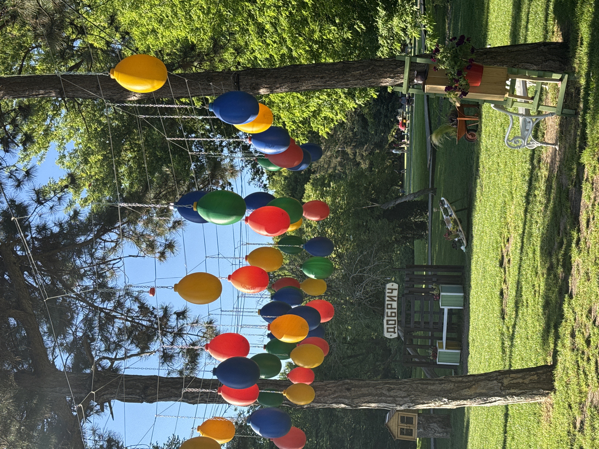 The image depicts a park in Dobrich, Bulgaria, featuring numerous colorful, bulbous decorations suspended from chains between trees. These decorations, in shades of yellow, red, blue, and green, create a festive atmosphere. In the background, a wooden sign with Cyrillic lettering that reads "ДОБРИЧ" is visible, identifying the location. A wooden bench with green and white accents is placed in front of a low wooden fence, and there are flower pots on either side of the bench. To the left of the bench, a small, rustic wooden structure resembling a miniature house is attached to a tree trunk. Further back, people are seen sitting at picnic tables, enjoying the outdoors. The ground is covered in lush green grass, and the scene is illuminated by sunlight filtering through the trees, casting dappled shadows. A white metal bench and a decorative ladder with flower pots are also present in the foreground.