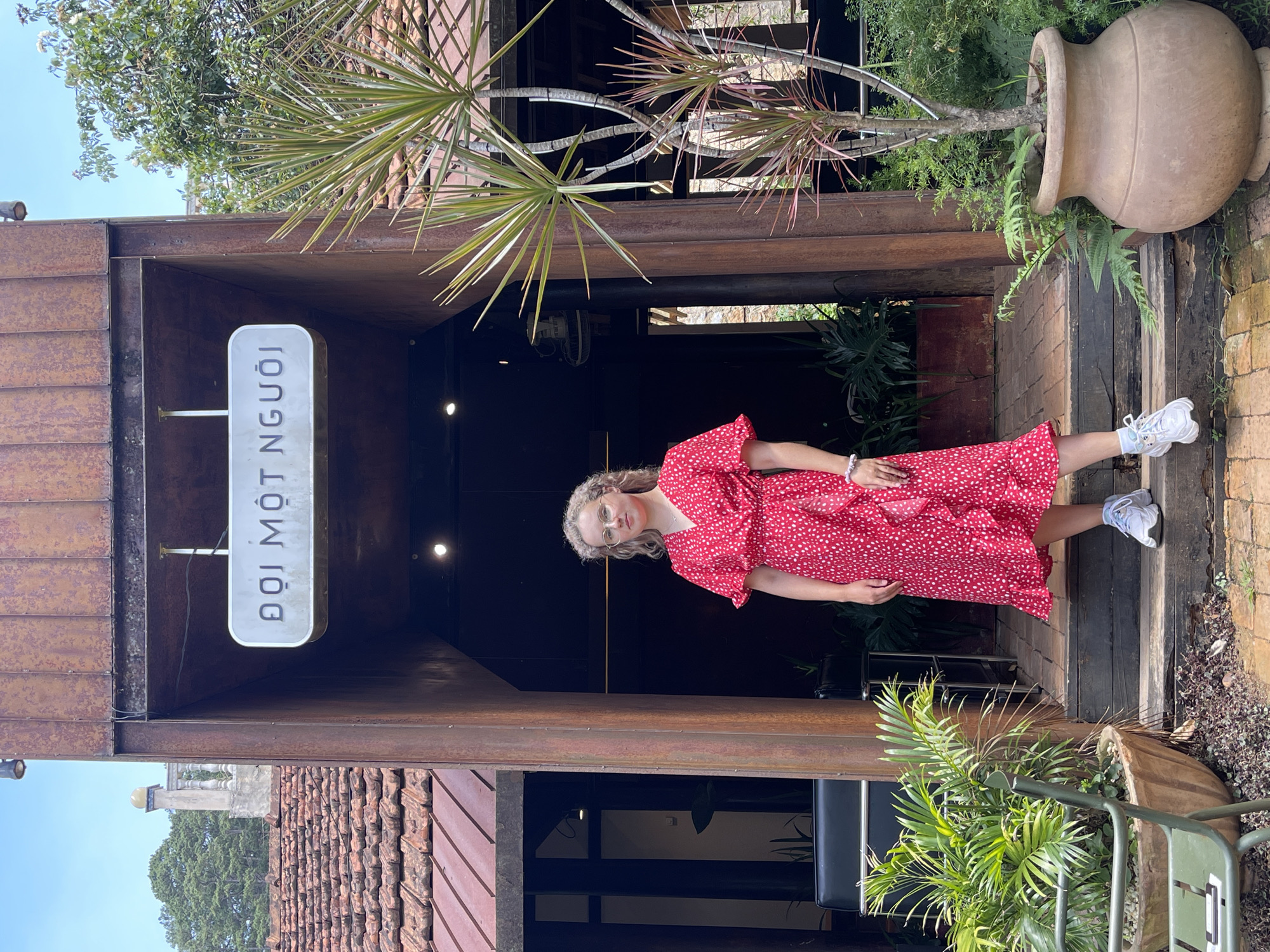 A vertical, medium shot captures a woman in a red dress standing in the entryway of a building with the Vietnamese text "ĐỢI MỘT NGƯỜI" on a sign. The building appears to be in Da Lat, Vietnam.
The woman, wearing glasses, is positioned slightly to the right of the frame. She has light skin and curly hair. She is wearing a short red dress with white polka dots. Her feet are clad in white athletic shoes.
The entrance has a weathered, rusty aesthetic. A large terracotta-colored pot is visible to the right, with a plant spilling out. To the left, a similar structure is seen with a brown-tiled roof.
The entryway leads into a dimly lit interior. The foliage of trees and plants is present, giving the impression of a garden setting. The sky is a clear blue in the top left corner of the image.