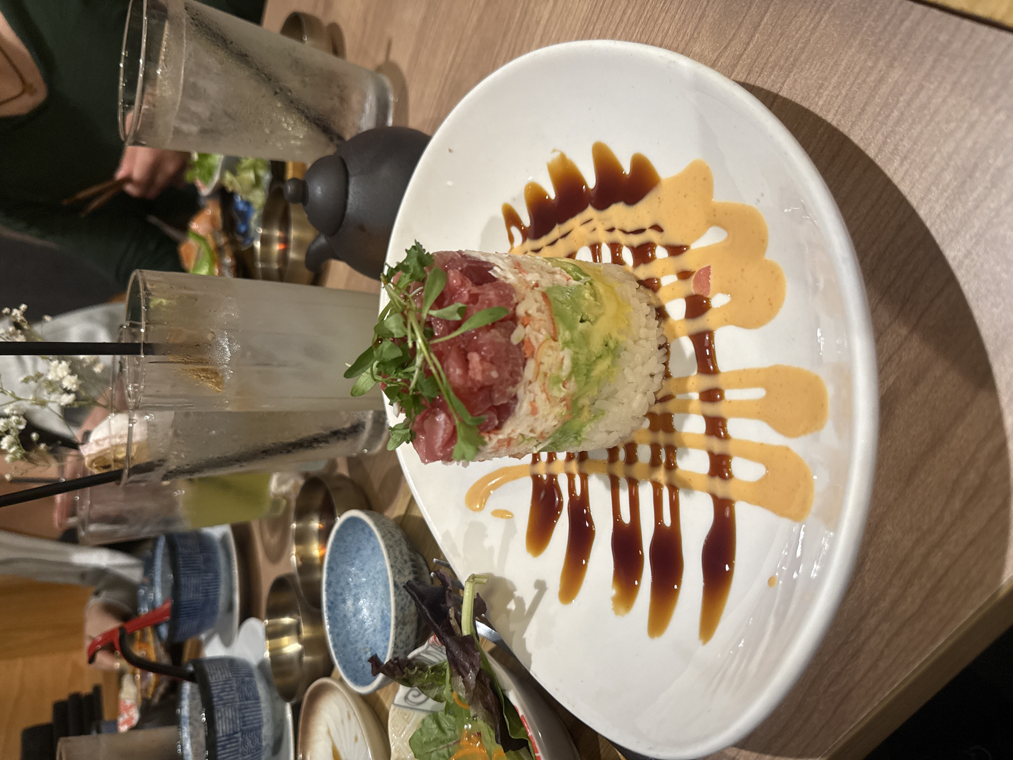 The image shows a plate of sushi on a wooden table. The sushi is cylindrical, with visible layers of rice, avocado, and tuna. It is topped with a small amount of greens. The plate is decorated with drizzles of brown and light orange sauce. In the background, there are glasses, a small teapot, and bowls. The image was taken at a restaurant in Southlake, United States of America.