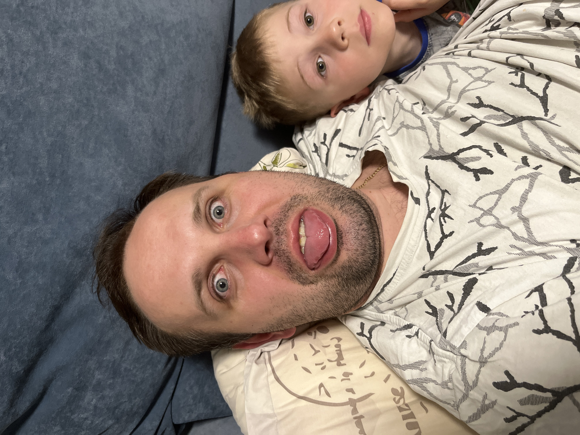A selfie photograph depicts an adult male lying on a bed or sofa, with a child positioned near his upper body. The man is looking directly at the camera with wide eyes and his tongue sticking out. He has short dark hair, a light complexion, and a beard. He is wearing a white t-shirt with a branch-like pattern in gray and black. A gold chain is visible around his neck. The child has light brown hair and is looking slightly off-camera. The setting appears to be indoors, possibly a home, with a blue upholstered surface visible on one side. The image does not provide specific details to confirm the location as Rusanovo, Russia.