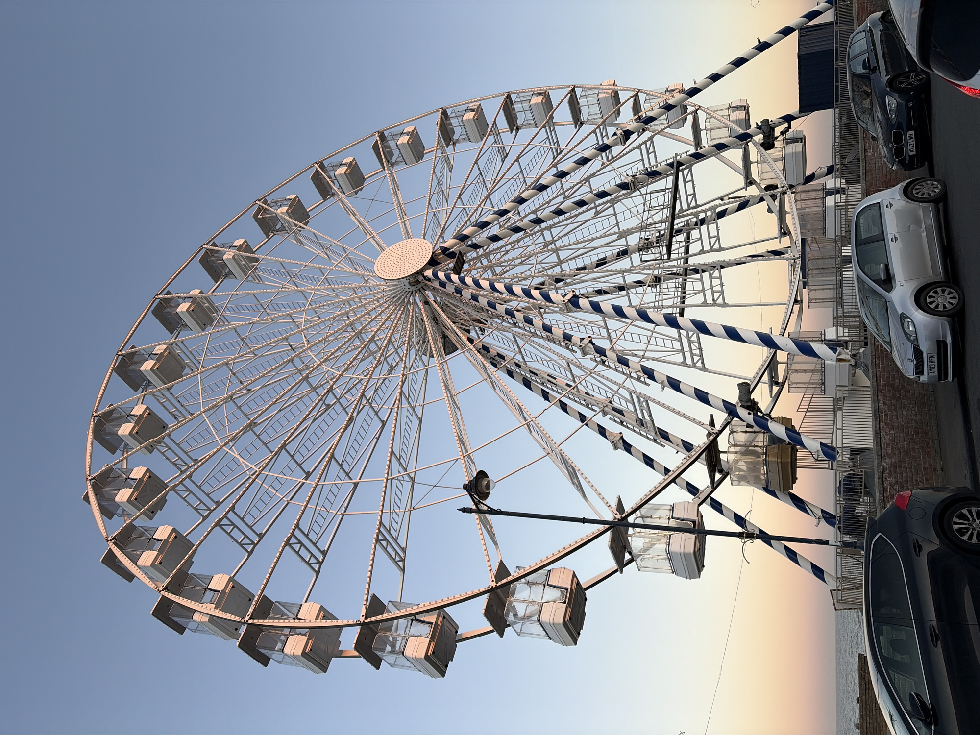 The image displays a large white Ferris wheel under a clear sky, captured from a low angle. The Ferris wheel, the primary subject, features numerous passenger gondolas and has several of its structural spokes wrapped in a distinctive blue and white striped pattern. A circular, textured hub is visible at the center of the wheel, and a light fixture is attached to one of its support beams.
The sky exhibits a gradient, transitioning from a light blue at the top to soft hues of orange, yellow, and pink towards the bottom right, suggesting either dawn or dusk.
To the right of the Ferris wheel, several cars are parked on a paved area, likely a street or promenade. A silver Fiat Punto with the UK license plate "AY63 HFM" is prominently visible, as is a dark blue BMW 1 Series with the UK license plate "NV12 LNN". Parts of other vehicles, including a dark-colored car in the foreground and a red car in the upper right corner, are also present. Beyond the parked cars, a low barrier or wall separates the paved area from the calm sea, which extends to the horizon. No people are explicitly visible, and no specific activity beyond the presence of the Ferris wheel and parked cars is taking place. The scene suggests a quiet moment at a coastal location, consistent with East Suffolk, United Kingdom.