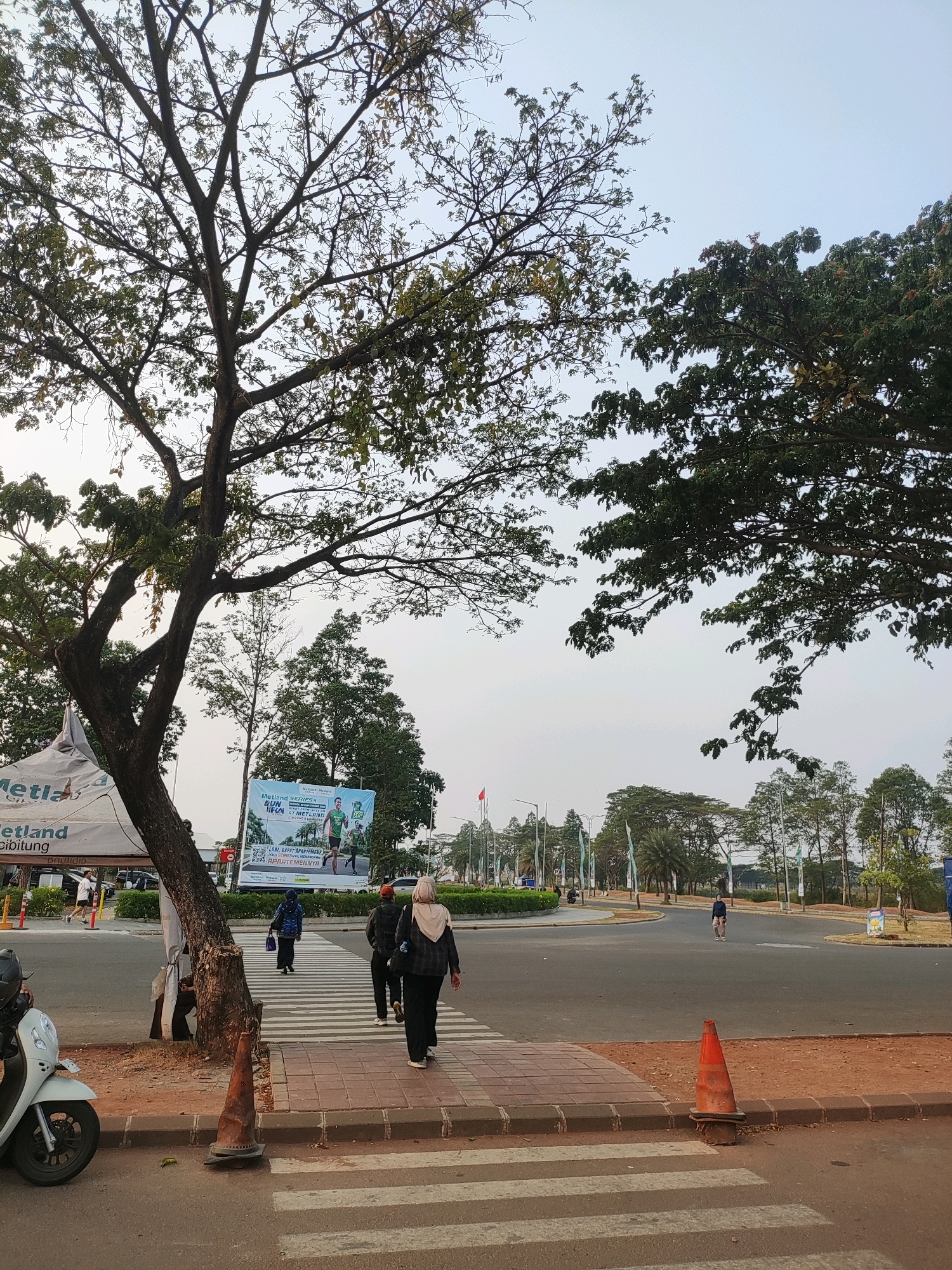 The image captures a scene in Telagamurni, Indonesia, featuring a pedestrian crossing on a road. In the foreground, a white scooter is parked to the left, and an orange traffic cone stands on the sidewalk near the crossing. Several people are walking across the zebra-striped pedestrian crossing, with some walking away from the viewer. A large tree with extensive branches dominates the left side of the frame, with its foliage partially obscuring the sky. In the background, a banner is visible, advertising a "Metland Series Run" event. The road appears to be a thoroughfare with multiple lanes, and there are trees and some buildings visible in the distance. The sky is overcast, suggesting a diffused natural light.