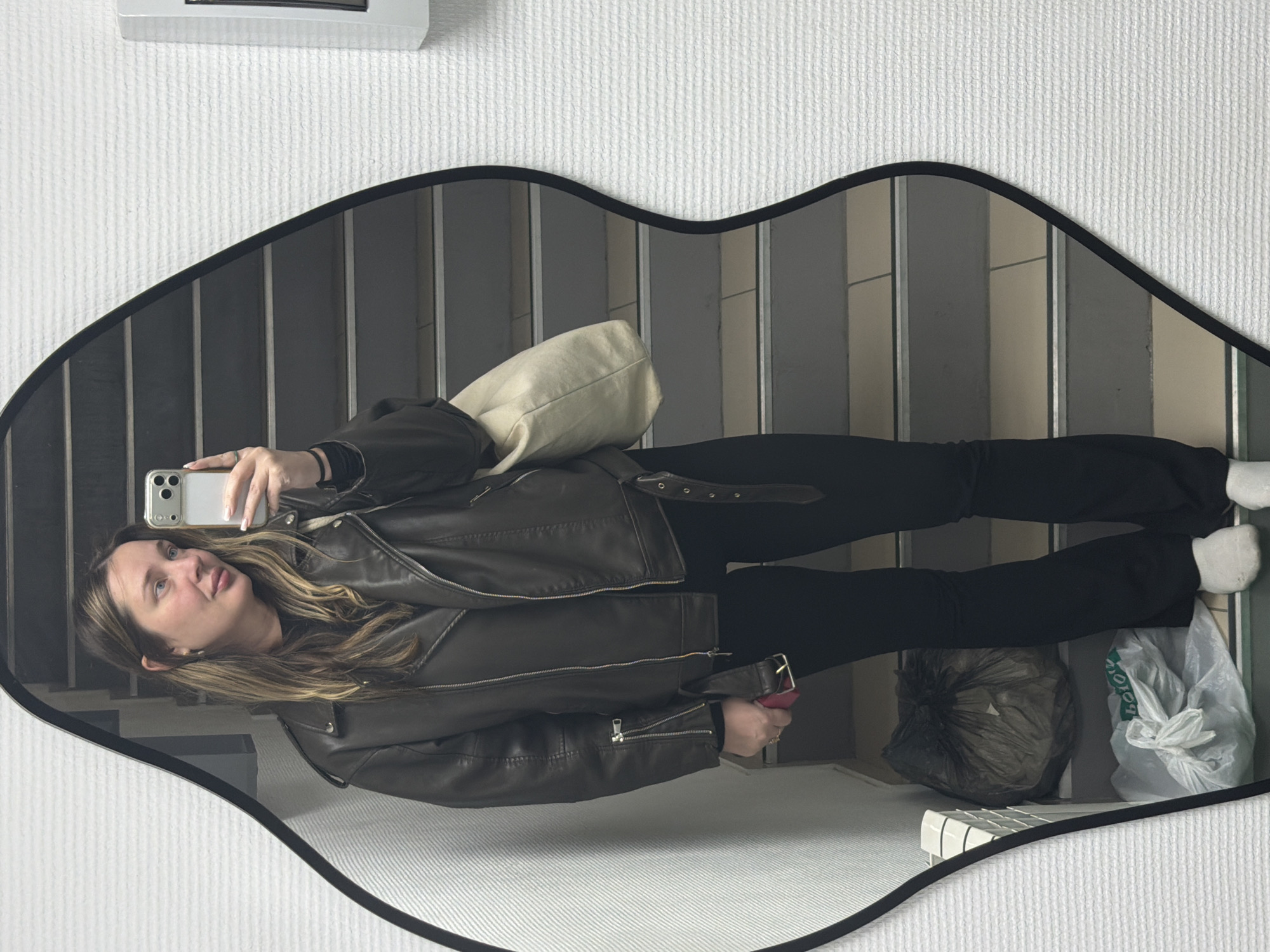 A young woman is taking a selfie in a lip-shaped mirror. She is wearing a brown leather jacket, black pants, and white socks. She is holding a white iPhone in her right hand. Her left hand is holding a red item. Behind her is a staircase. There is a white tote bag slung over her left shoulder. Two garbage bags are visible to the right and below her. The mirror is hanging on a white wall, with a white air conditioner unit visible above the mirror. The reflection in the mirror suggests the scene is indoors, possibly near a stairwell. There is no information indicating that this image was taken in Chaikovsky, Russia.