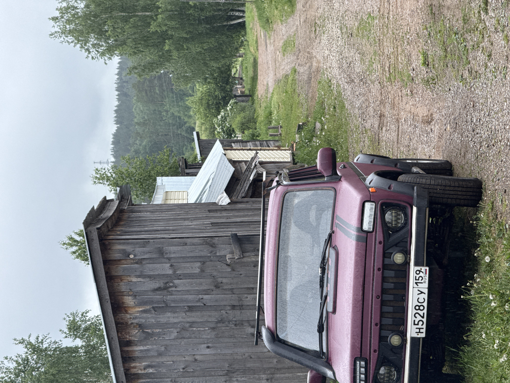 The image depicts a burgundy Lada Niva SUV parked adjacent to a weathered wooden building, possibly a shed or house, on a gravel road in Прикамский, Russia. The car's license plate reads "H528CY 159 RUS". Vegetation, including grass and trees, surrounds the area. A forest is visible in the distance under a cloudy sky, suggesting a rural or semi-rural setting. The scene appears to be taken on a damp day.