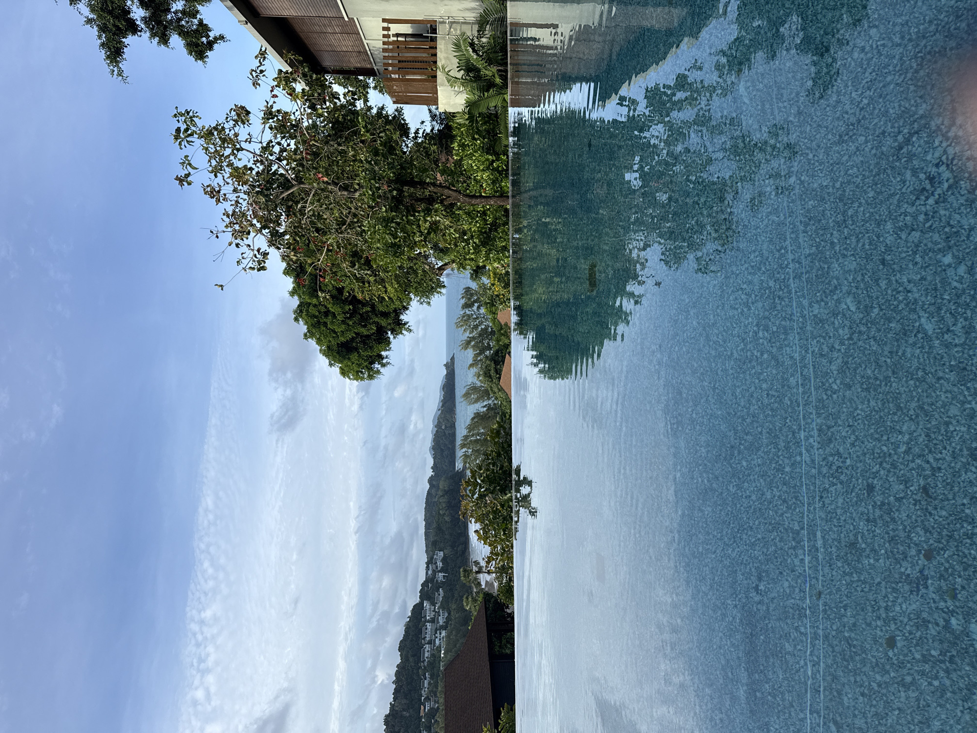 An infinity pool is in the foreground, reflecting the sky and trees. Beyond the pool, a verdant coastline stretches across the horizon, dotted with buildings and a beach. The sky is cloudy with patches of blue. The location is Sakhu, Thailand.