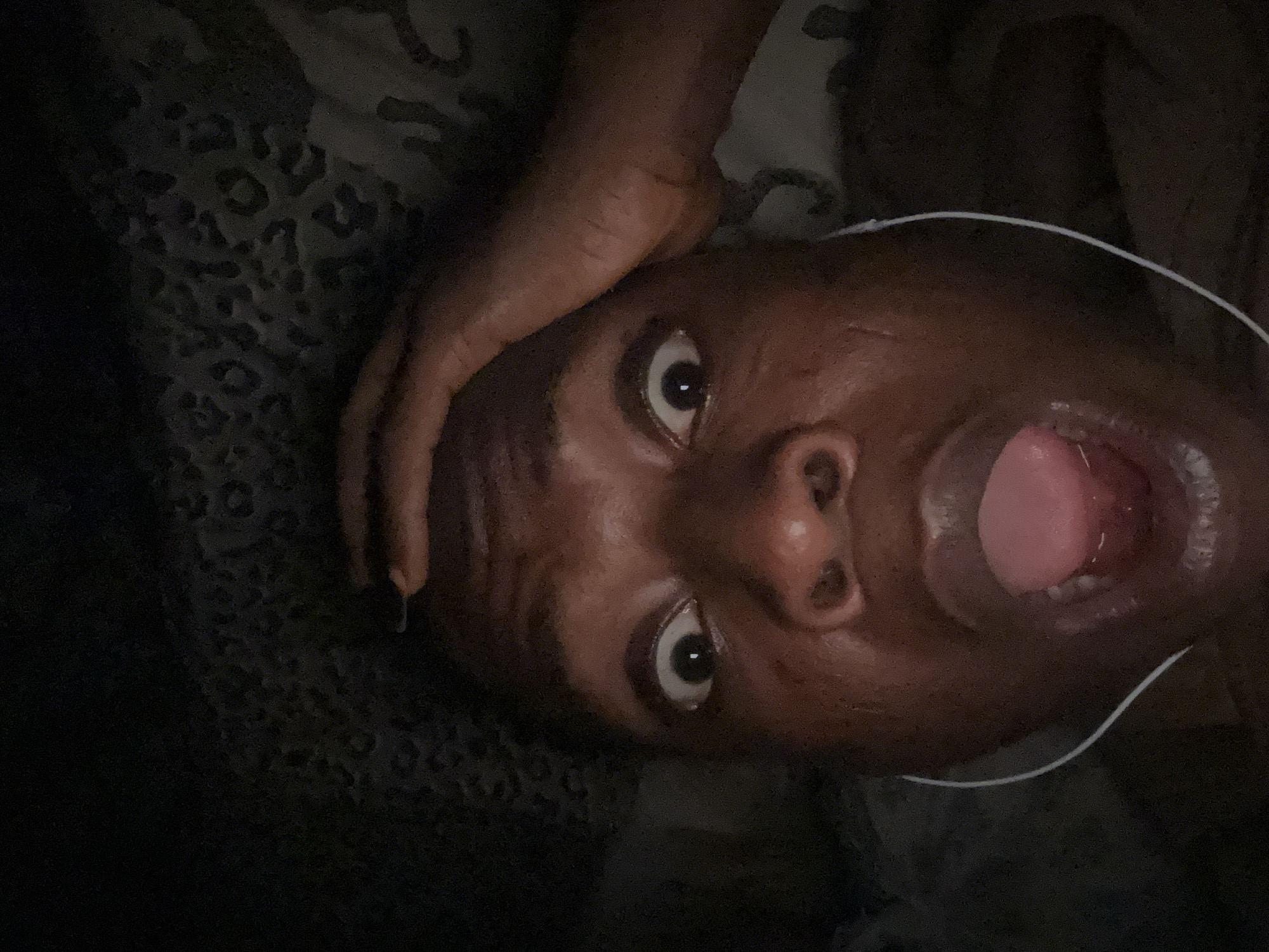 A close-up, low-angle shot depicts a person with dark skin lying down, their head tilted upwards. The person's eyes are wide and their mouth is open in an expression of surprise or shock. A hand rests on their forehead, with fingernails appearing to be painted dark. White headphones are partially visible near the person's right ear. The background consists of a dark, patterned fabric. The location is Unknown, Unknown.