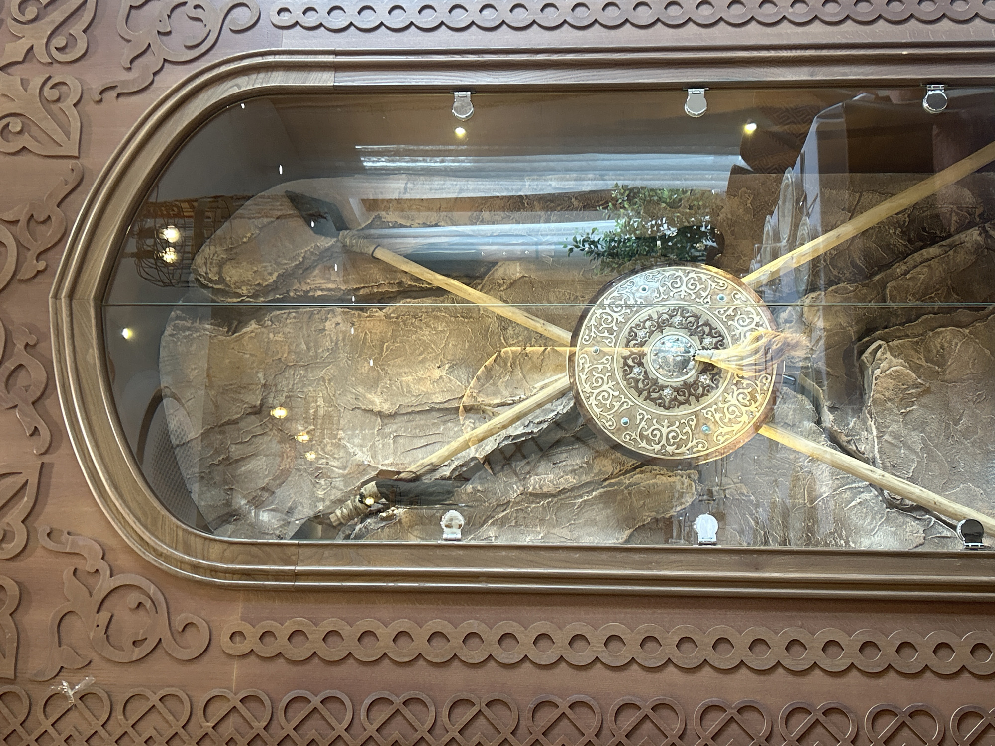 The image shows a display case in a building, possibly a museum or a traditional dwelling. The display case is tall and arched, with ornate wooden carvings on the frame, featuring geometric and intertwining patterns. Inside the case, a large, decorative shield or gong with intricate golden patterns and a tasseled ornament is prominently displayed, leaning against a faux rock formation. Two wooden poles support the shield. The background of the display includes a dim interior with what appears to be a table set for a meal visible through the glass, and sheer curtains in the distance. The lighting within the display case is subdued, with some visible light fixtures. The exterior of the display case has metallic handles. The overall impression is one of cultural heritage preservation and display. This display case is likely located at the "Nazarbayev Center" in Almaty, Kazakhstan, an architectural complex dedicated to preserving and promoting Kazakhstan's cultural heritage, often featuring traditional artifacts and art.