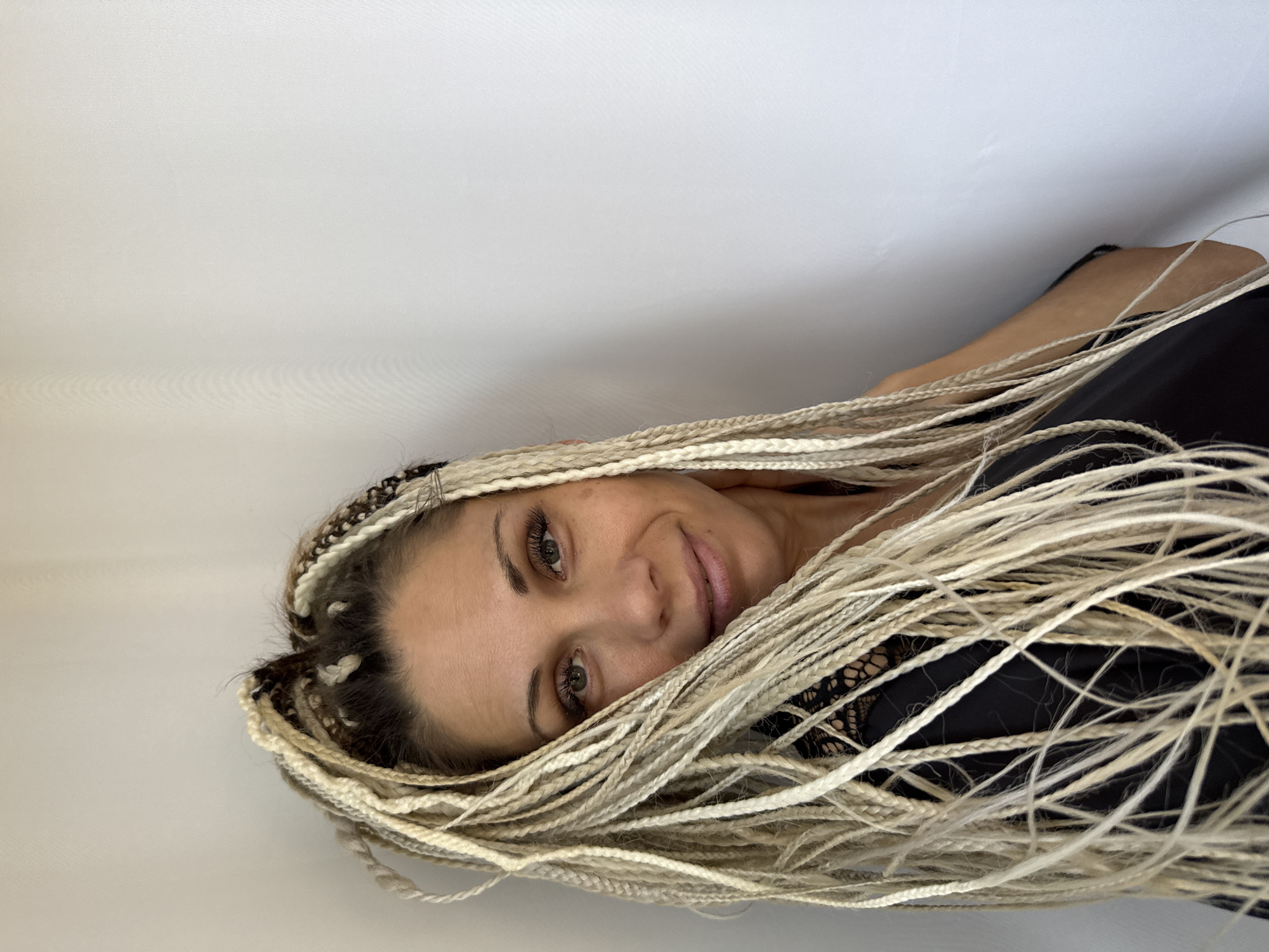 A portrait of a woman with blonde box braids, wearing a black top with lace detail, is shown against a plain white wall. She is looking towards the viewer with a slight smile. The background is a plain, textured white wall. The image is cropped at chest level, with the woman's face and hair dominating the frame. The scene does not depict any specific urban location.