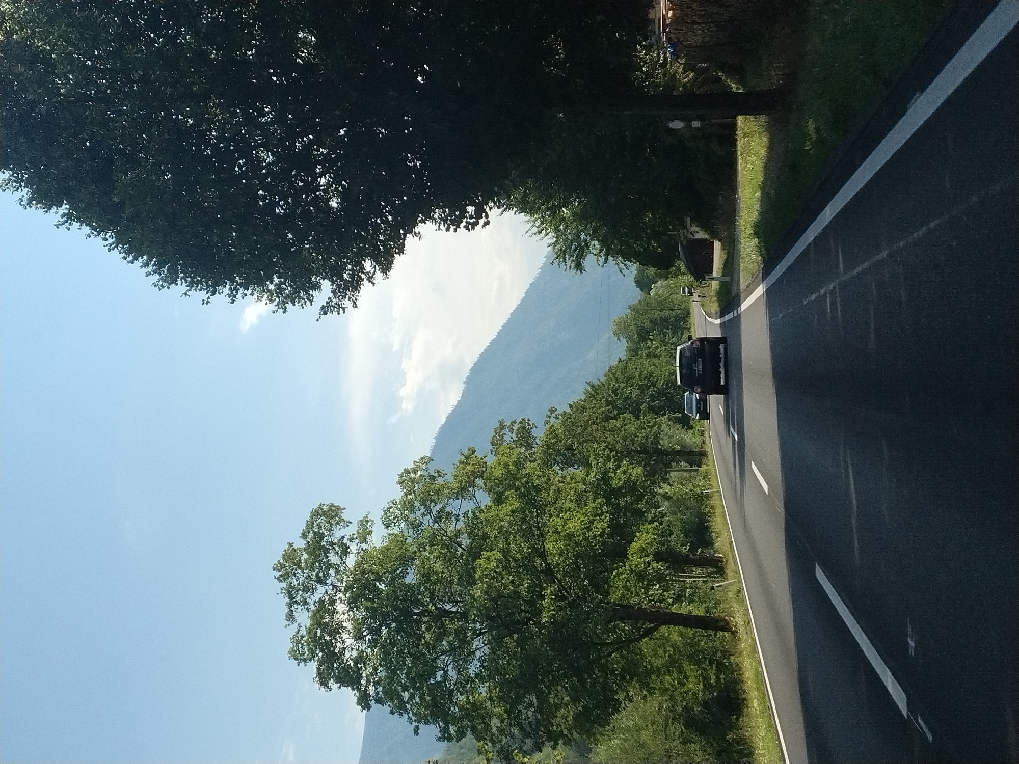 A dark-colored car drives on a two-lane asphalt road with white lane markings. The road curves to the right, flanked by trees and grass. In the background, a large, green mountain range is visible under a clear blue sky with a few wispy clouds. The scene appears to be the Salzkammergut region of Austria, possibly near Ebensee, given the mountainous terrain and dense vegetation. The car's headlights are on.