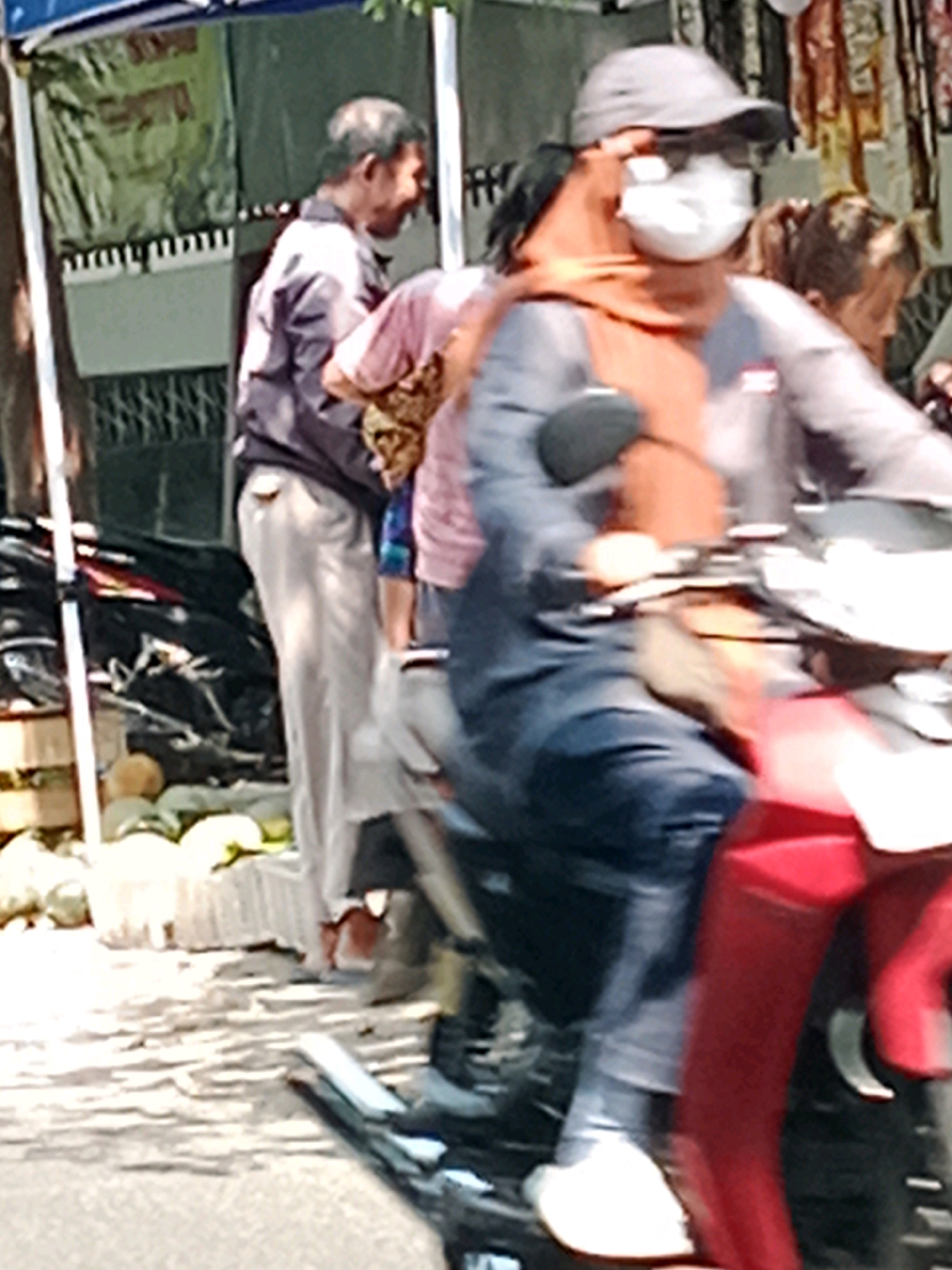 In Banaran, Indonesia, on a paved road, a person wearing a dark blue jacket and blue jeans sits on a red motorcycle. They are wearing a grey baseball cap, sunglasses, and a white face mask. To the left of the motorcycle, a man in a grey jacket and light-colored pants stands. Behind the seated person, another individual in a pink shirt is visible. The background consists of a stall with produce, possibly fruit or vegetables, including several round, light-colored items. The scene appears to be outdoors during daylight.