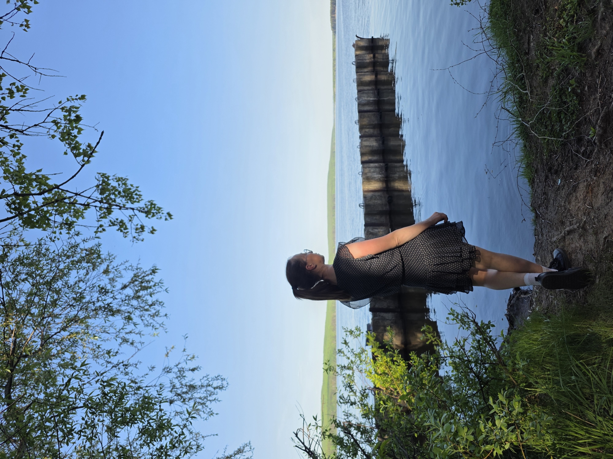 A woman stands beside a body of water. She wears a dark-colored dress with a light pattern, white socks, and dark shoes. Her hair is pulled back in a ponytail. She looks out at the water. There is a wooden barrier in the water. Green trees and bushes are visible. The sky is blue. This scene is in the city of Chaikovsky, Russia.
