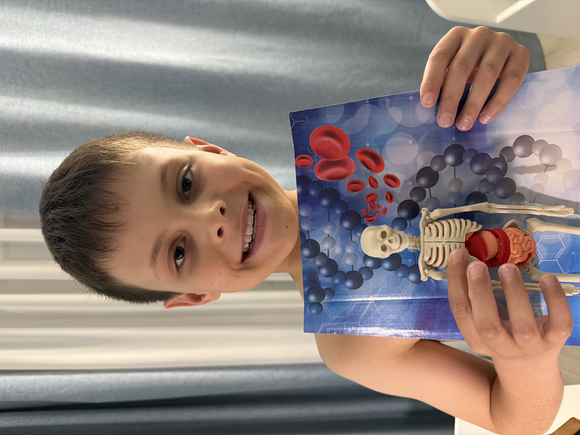 A young boy with short brown hair and fair skin smiles at the camera while holding up a piece of paper with a diagram of the human circulatory system and skeleton. The diagram features red blood cells, blue molecular structures, a skeleton, and internal organs. The boy's hands are visible, holding the paper. The background includes a gray and white curtain. The location, Votkinsk, Russia, is not discernable from the visual details provided in the image.