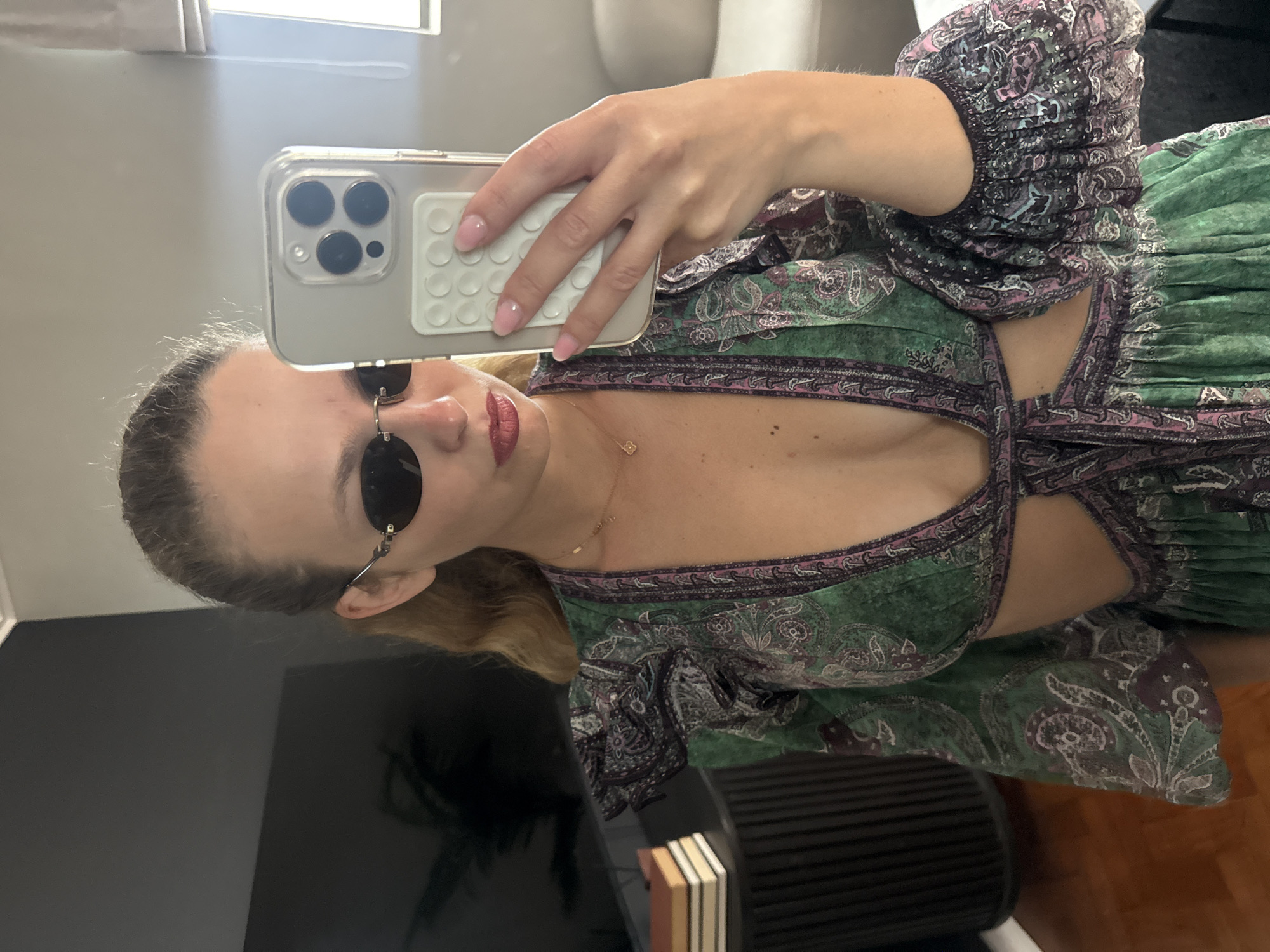 A woman is taking a selfie using an iPhone. She is wearing round sunglasses and a patterned green dress with a deep neckline. The background shows a glimpse of a room interior with a dark, reflective surface (possibly a TV screen), a window with white curtains, a light-colored wall, and a portion of a chair. The location in the city of Cape Town, South Africa cannot be determined from the provided image.