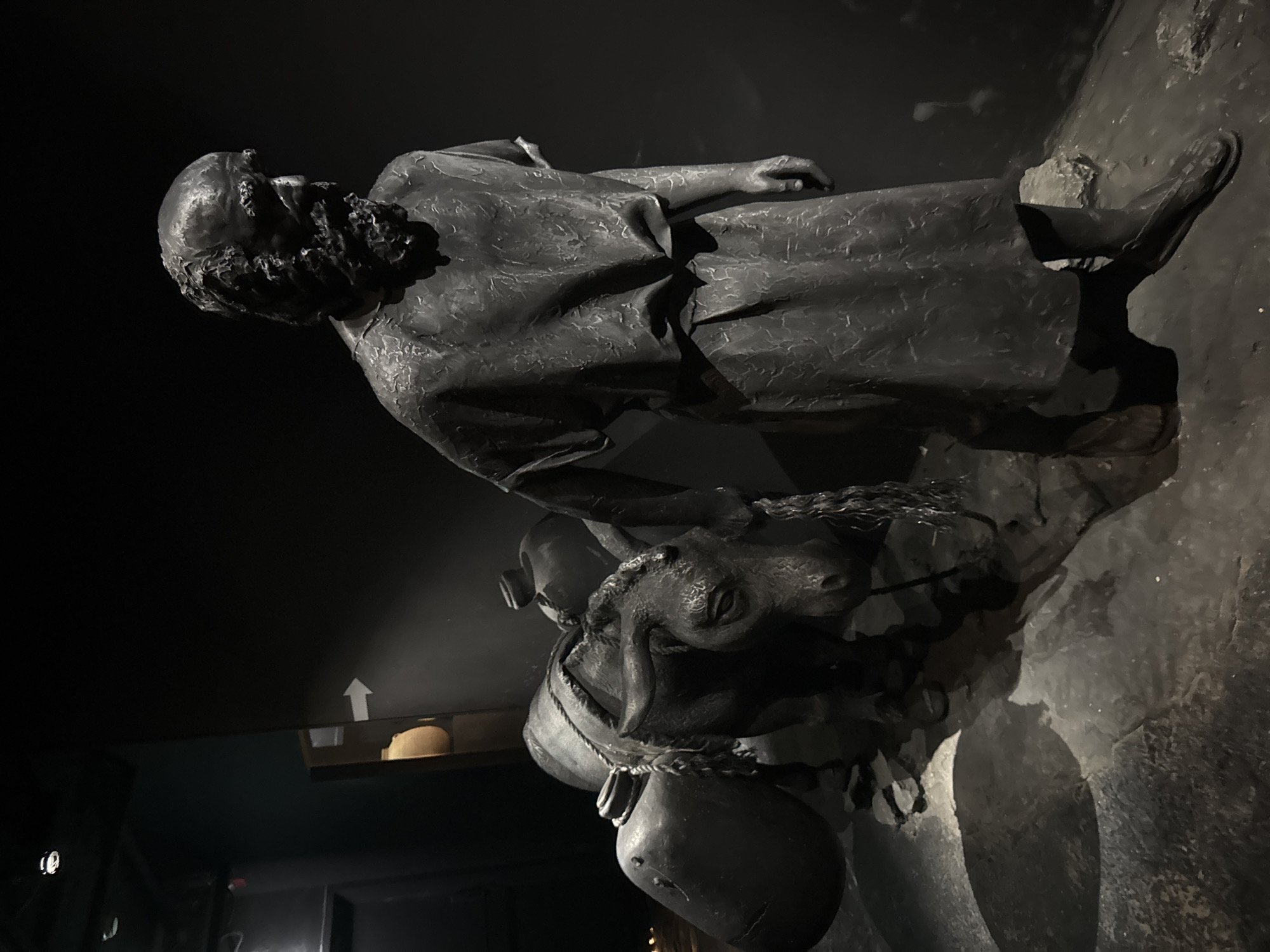 In the city of Tamar Regional Council, Israel, a dark, atmospheric display features two life-sized, monochromatic sculptures. The primary subject is a man with a rough-hewn beard and hair, wearing a simple, draped garment and sandals. He is shown in profile, looking downwards and to his right, with his right hand resting on the reins of a donkey. The donkey, also a sculpture, stands to the man's left, laden with two large, bulbous sacks on its back, secured by a rope harness. The donkey's head is slightly bowed, and it appears to be a dark, stone-like material, mirroring the man's sculpture.
The sculptures are positioned on a textured, uneven surface that resembles stone or compacted earth, with loose debris scattered around their feet. The lighting is dim and directional, casting strong shadows that emphasize the textures of the sculptures and the ruggedness of the ground. To the left of the man and donkey, in the background, a portion of a wall is visible, with a prominent white arrow pointing to the right, suggesting a directional marker within the exhibition space. Further in the background, a hint of a curved, ochre-colored pot can be seen on a shelf. The overall environment is one of subdued lighting and a focus on the historical or symbolic representation of the figures.