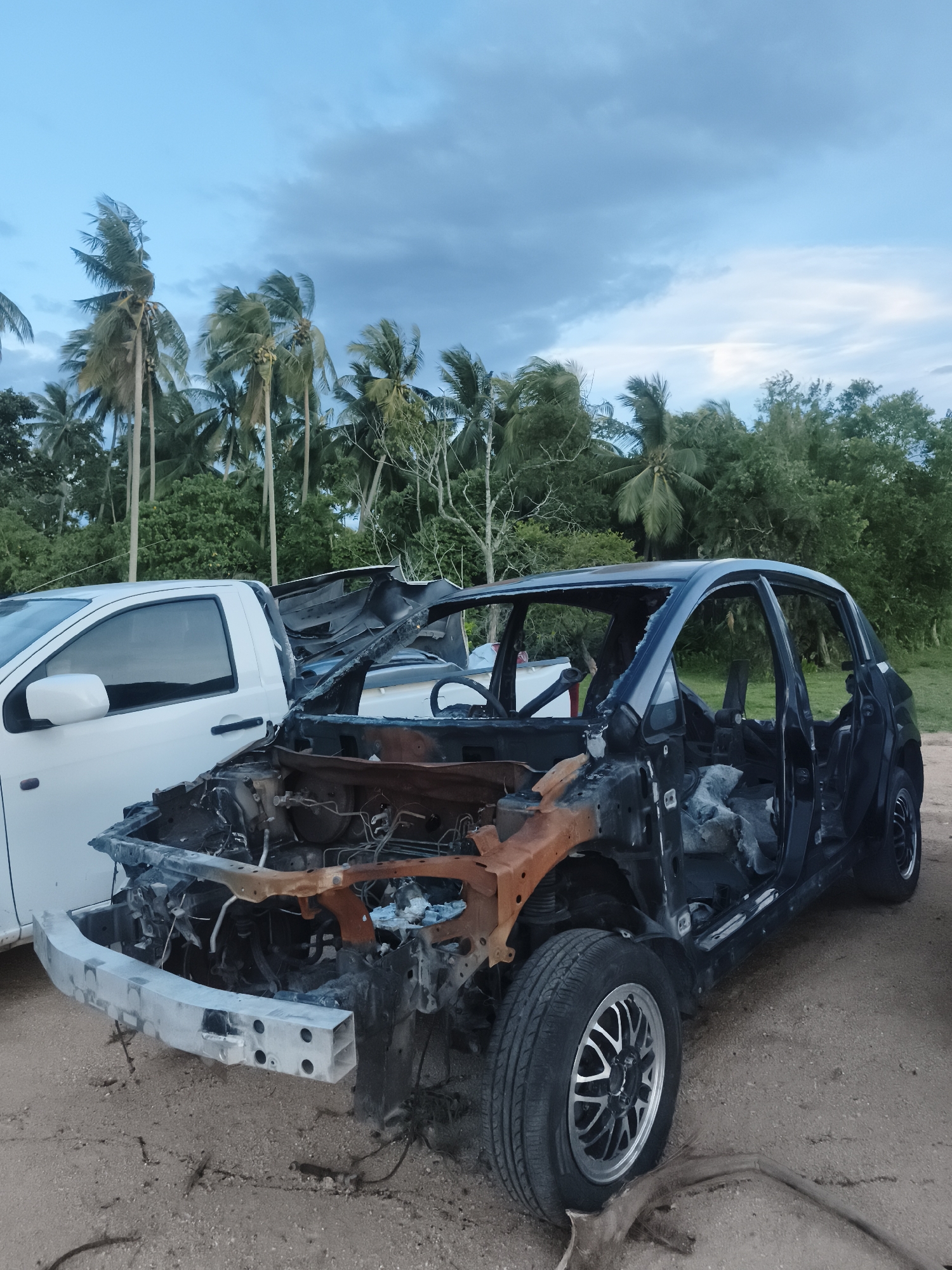 The image depicts a severely damaged, likely burnt-out car chassis in an outdoor setting. The car's body is largely disintegrated, exposing the rusted engine compartment and interior. Only the frame, wheels, and some structural elements remain. A white pickup truck is parked closely to the left of the car chassis. The background includes palm trees and other lush vegetation, typical of a tropical environment. The sky is overcast. The scene is located in Baan Mae Nam, Thailand. The ground appears to be sandy or dusty. No people are visible.