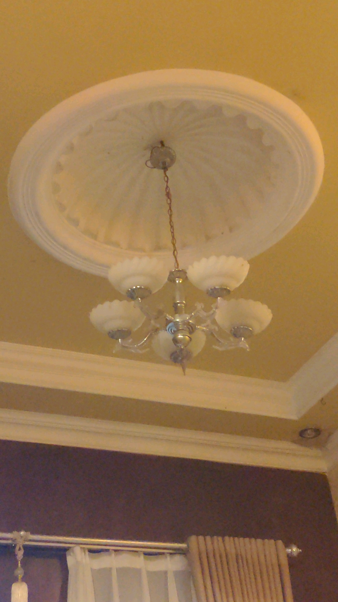 The image shows a chandelier hanging from a decorative ceiling medallion. The medallion is circular with raised, textured patterns radiating from the center. The chandelier has a metallic finish and multiple frosted glass shades. Below the ceiling, there are layers of molding and a dark purple wall. A curtain rod with white curtains and beige curtains is visible at the bottom of the frame. The scene appears to be an indoor setting, likely a room in a house or a building in Tembarak, Indonesia. There are no people or animals present in the image.