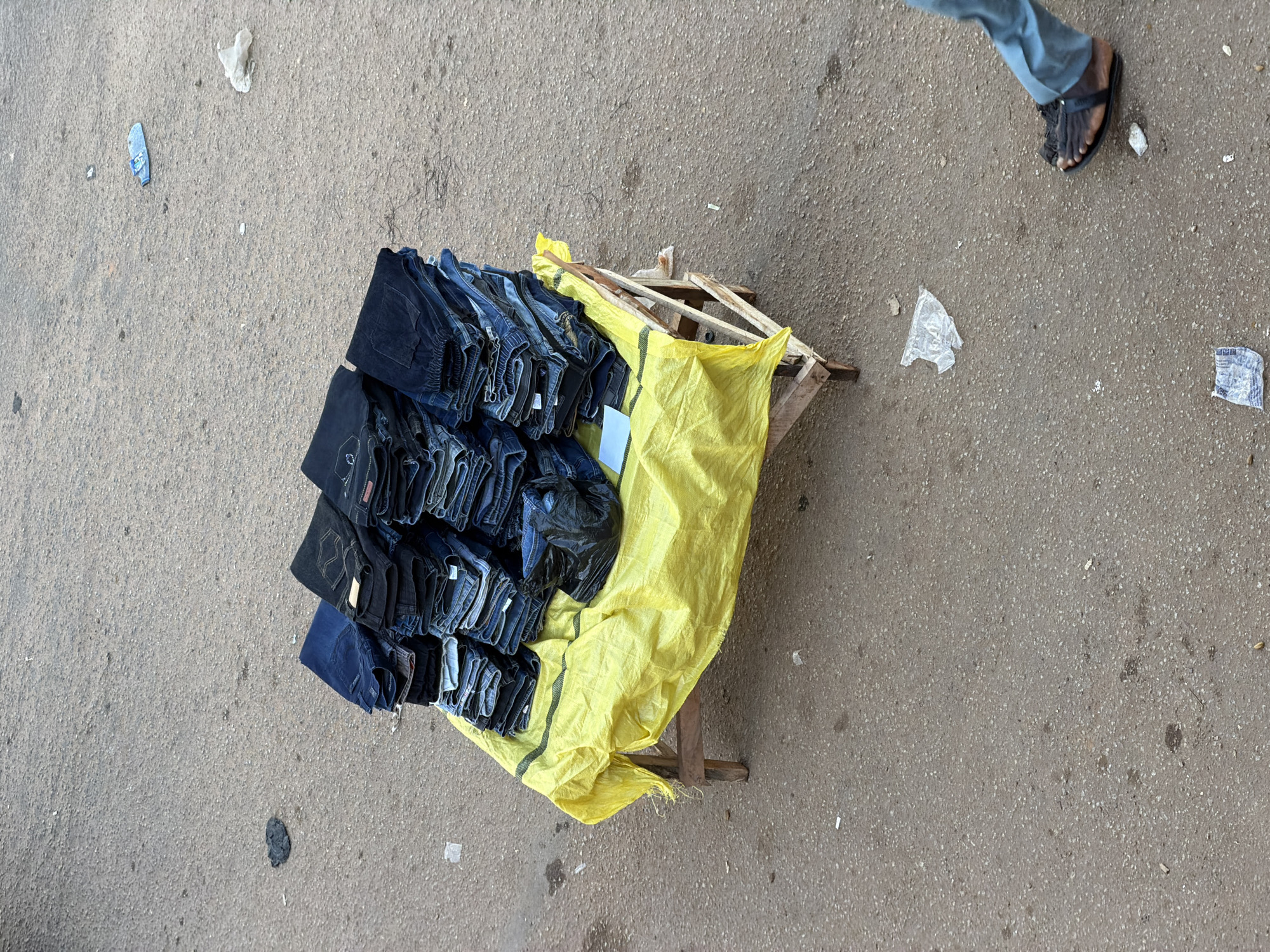 In Bukuru, Nigeria, a market scene unfolds with a wooden stand draped in bright yellow material, showcasing stacks of folded blue jeans. The jeans are neatly piled, some with brand labels visible. Next to the jeans, a black plastic bag contains more denim items. The ground is paved with asphalt, showing signs of wear and scattered debris, including discarded plastic bottles, wrappers, and a dark, amorphous lump. In the lower right quadrant, a person's legs and feet are visible, clad in blue trousers and dark sandals, indicating movement. The overall impression is a typical street market setting.