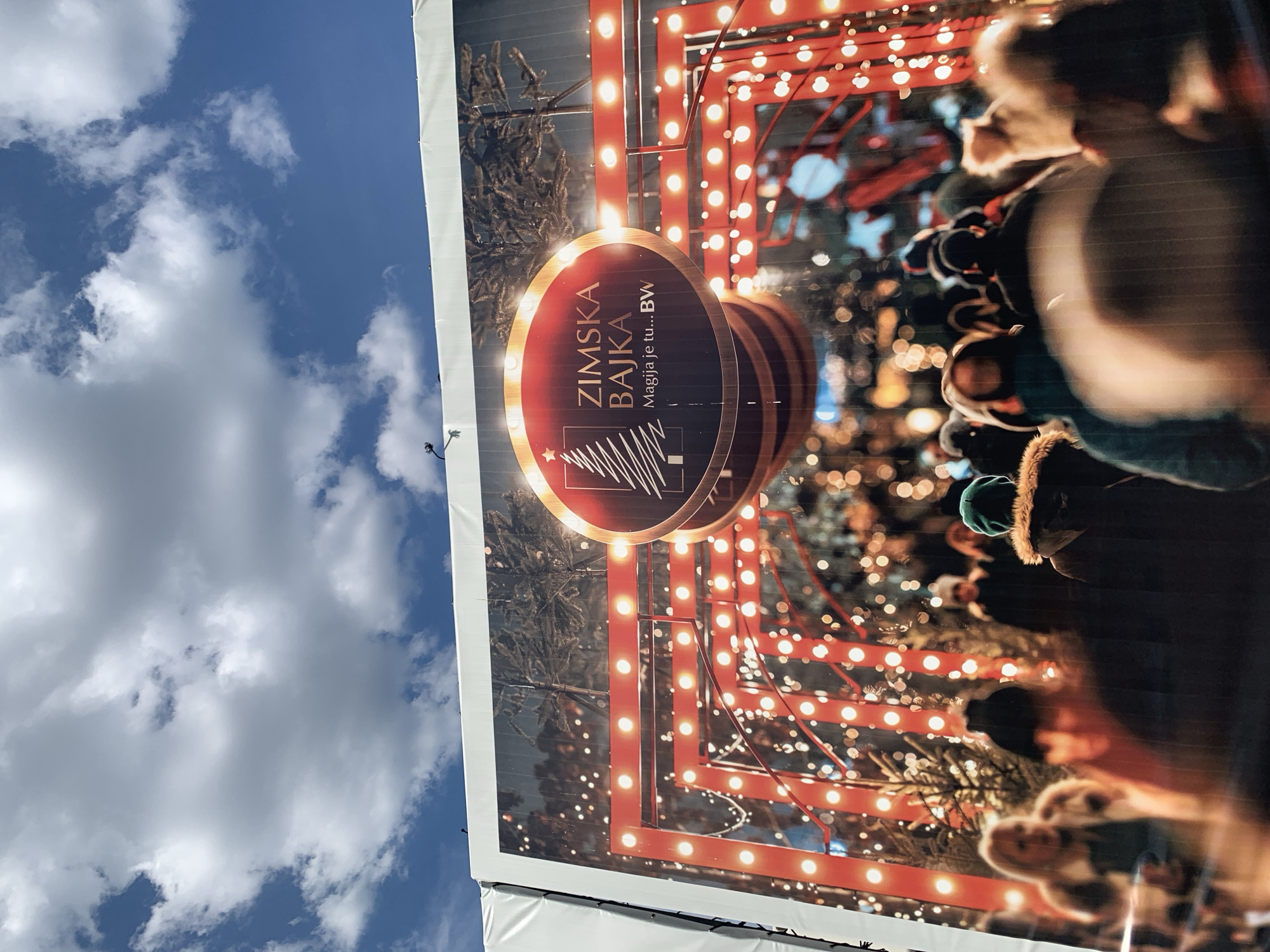 The image captures a large outdoor advertisement in Belgrade, Serbia. The ad promotes "Zimska Bajka," a winter-themed event, featuring the text "Magija je tu...BW". The display incorporates a design of a Christmas tree and festive lights. In the background, a crowd of people can be seen, suggesting the event is well-attended. Above the advertisement, the sky is visible with scattered white clouds against a blue backdrop.