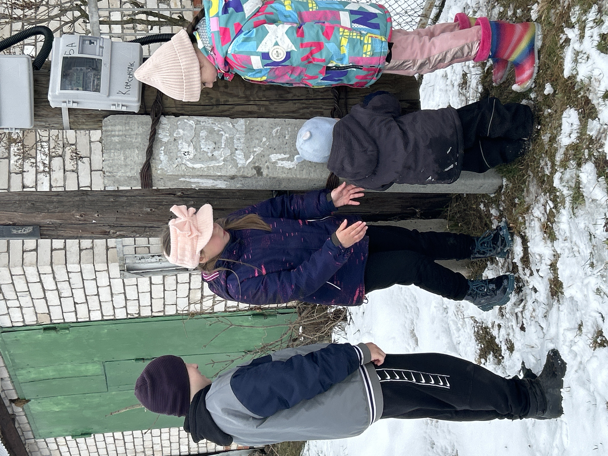 Four children are lying on a wooden structure outdoors in what appears to be a snowy environment. The location is in the city of Chaikovsky, Russia. The children are wearing winter clothing, including hats, jackets, pants, and boots. The structure they are lying on is made of wood and concrete, and it is attached to a building with a green door and brick walls. There is a utility box attached to the structure. One child is wearing a pink blindfold.