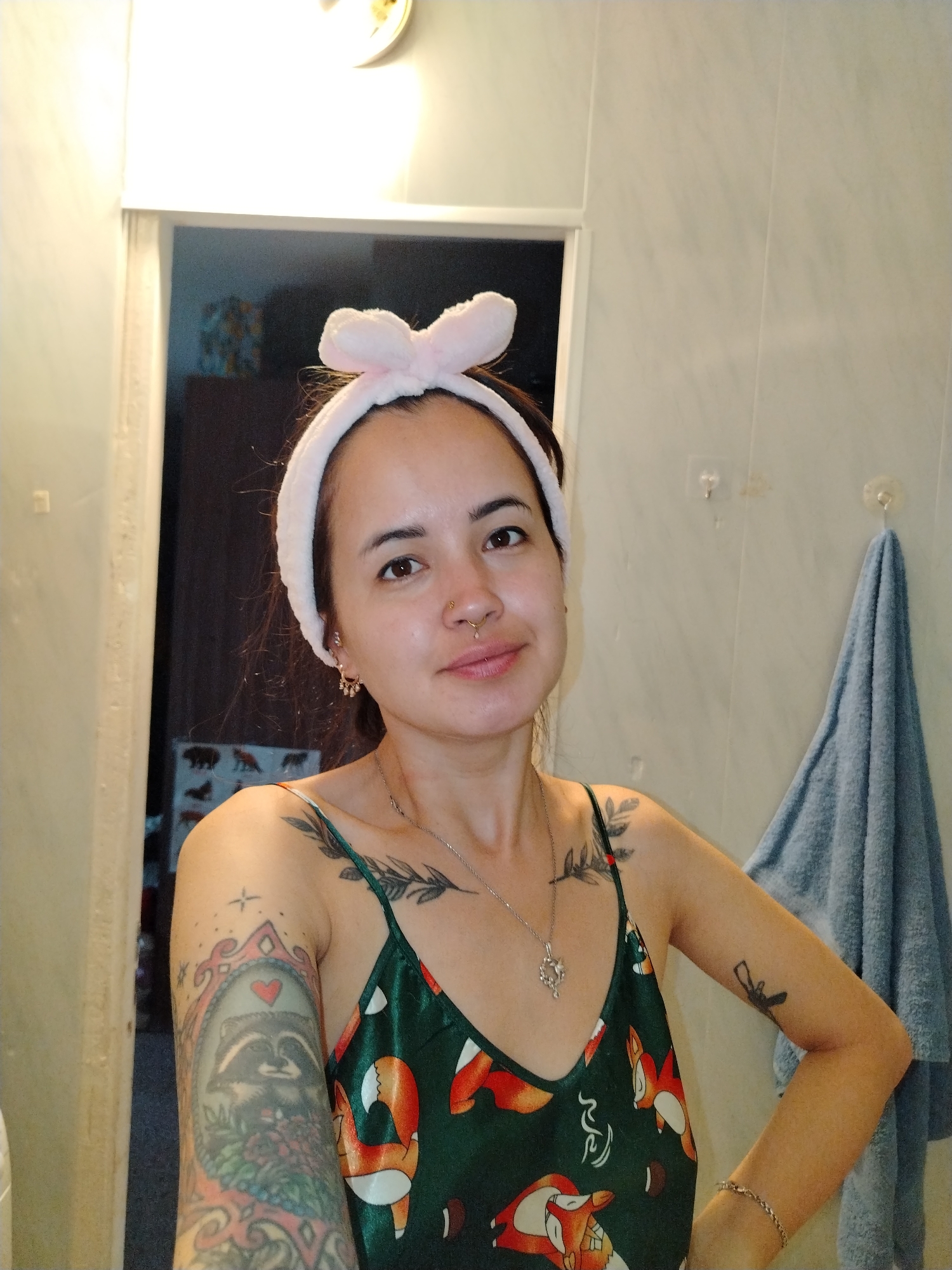 A young woman with a fluffy pink headband with rabbit ears is posing for a selfie in a bathroom. She is wearing a green camisole with a fox pattern. Her hair is dark brown and pulled back. She has a nose ring and earrings. She has tattoos on her left arm, shoulder, and chest. A light blue towel is hanging on the wall behind her. The bathroom has tiled walls.