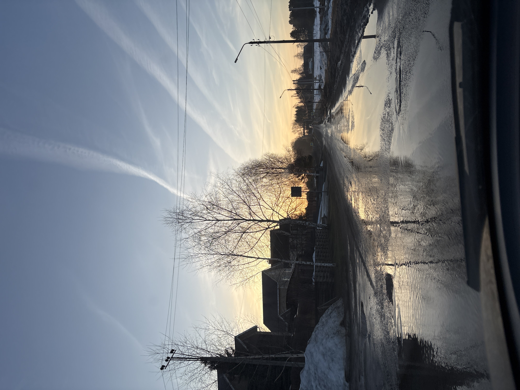 The image shows a street scene in Прикамский, Russia at either sunrise or sunset. The road is wet and reflective, indicating recent rain or melting snow. There are puddles on the road reflecting the sky. Buildings line the street, some with snow piled next to them. Bare trees are silhouetted against the sky. Power lines run along the side of the road. Contrails are visible in the sky.
