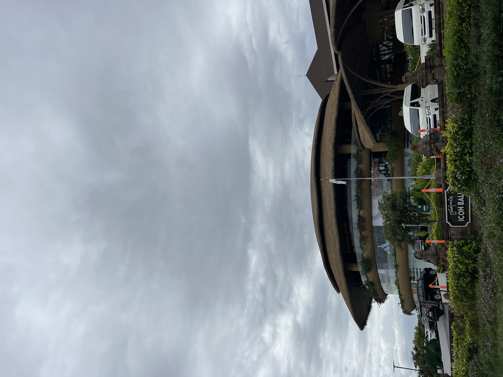The image shows the exterior of the "Icon Bali" shopping mall in Denpasar, Indonesia, under a cloudy sky. The building features a distinctive circular design with a thatched roof and glass facades. White vans are parked in front of the entrance, and a black SUV is visible in the parking lot. Landscaping with lush greenery and flowering plants surrounds the building, with a traditional Balinese stone wall in the foreground. A flagpole with an Indonesian flag is also present.