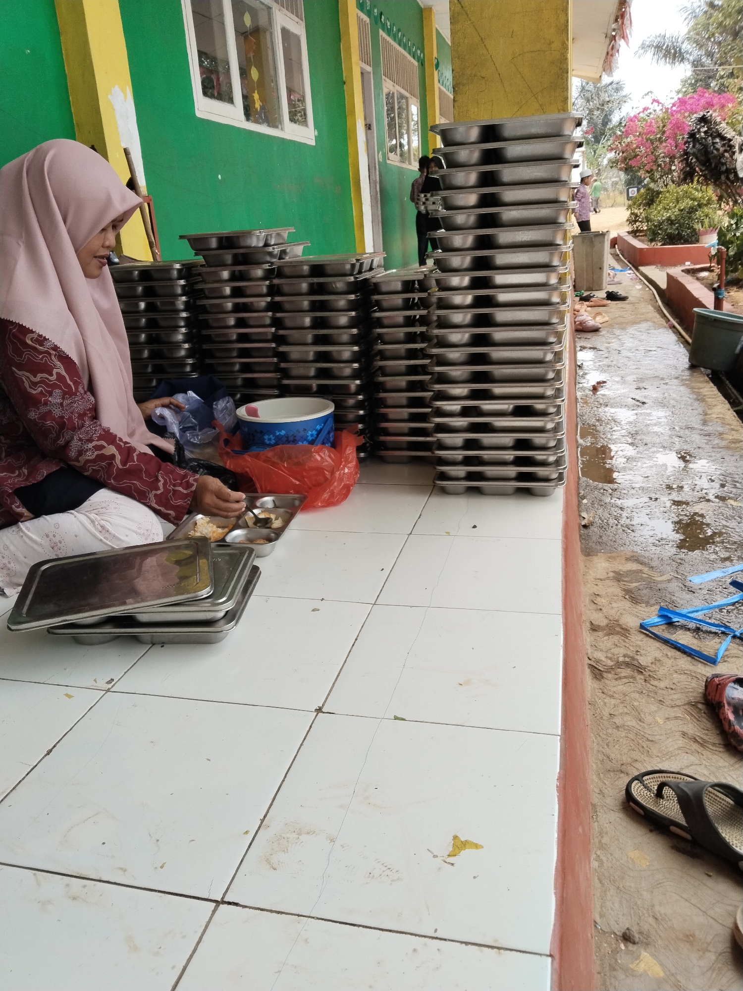 The image depicts an outdoor scene with a woman wearing a pink hijab and a maroon patterned shirt sitting on a tiled floor. She is in the process of sorting or preparing food in a metal tray, which contains several compartments with food items. A large stack of metal serving trays is visible to her right, and a blue and white patterned bowl sits in a red plastic bag nearby. The background shows a green and yellow building with windows, and beyond that, trees and flowering plants. To the right of the tiled area, there is a dirt path with some puddles of water, as well as a pair of black flip-flops. The overall setting appears to be an outdoor area adjacent to a building, possibly a school or a community center, in Serang, Indonesia.