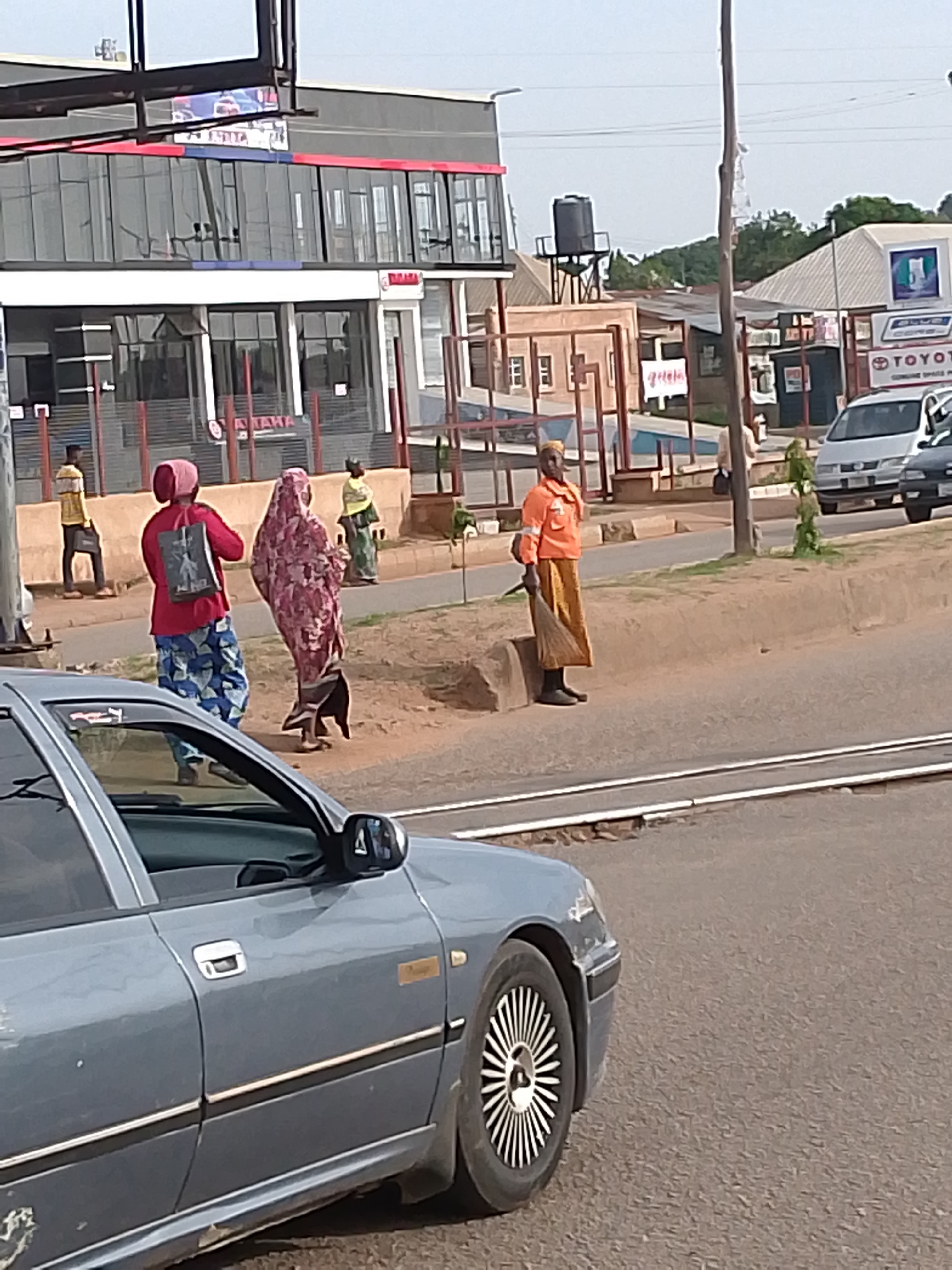 A street scene in Kuru Dungus, Nigeria. In the foreground, the front passenger side of a blue sedan is visible. Beyond the car, on the sidewalk next to a road, a person dressed in an orange long-sleeved shirt and a long yellow skirt stands holding a broom. Further down the sidewalk, three individuals are walking away from the camera. Two are women wearing headscarves and long patterned dresses, and the third is a man in a yellow shirt and black pants. Behind them, a modern building with large glass windows and red trim is situated. In the background, other buildings, trees, and a water tower are visible under a hazy sky. The road appears to have train tracks running across it.