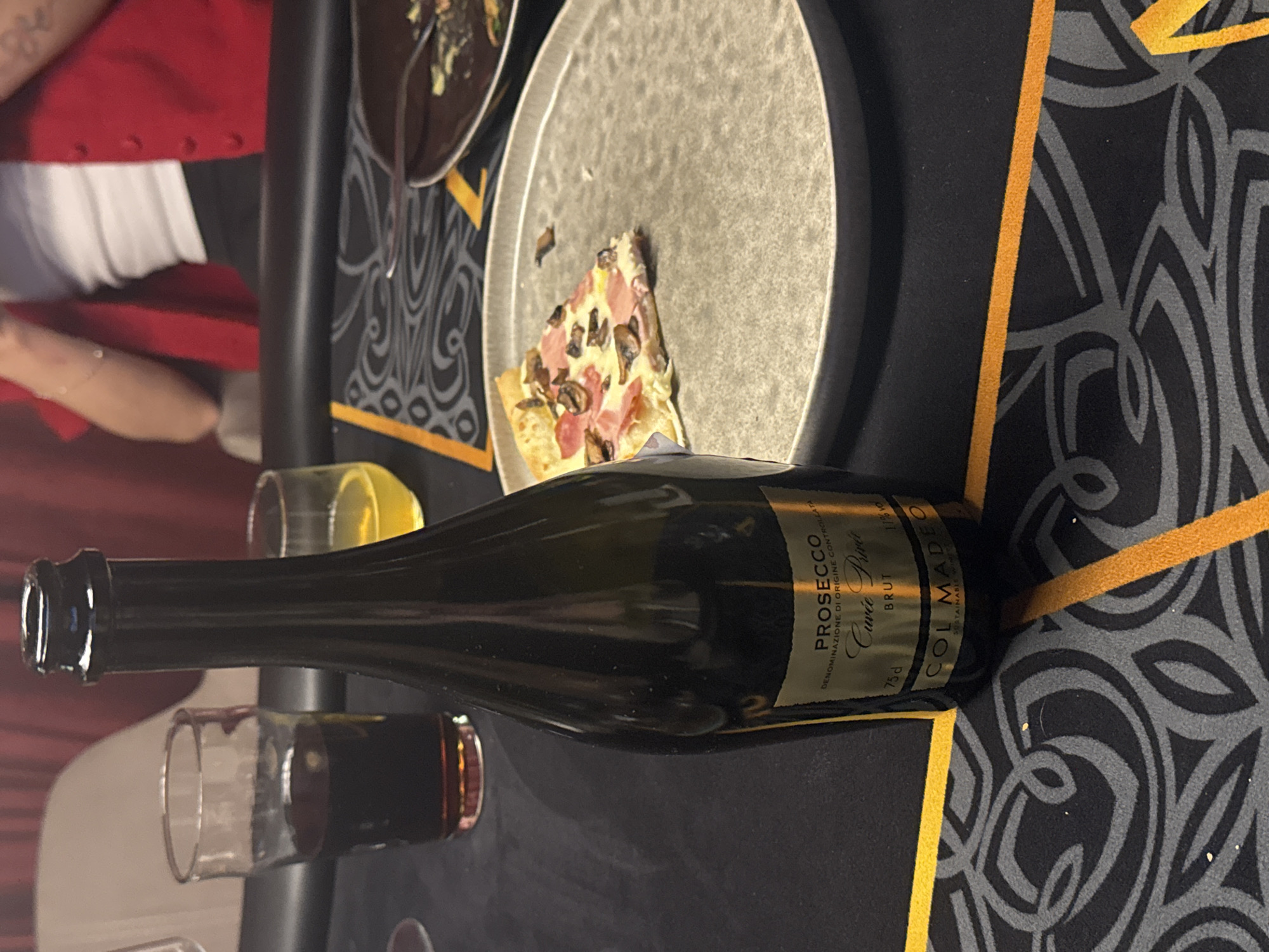 The image shows a table setting with a partially eaten slice of pizza on a plate. There is also a bottle of Prosecco Cuvée Privée Brut wine and two glasses, one with a clear liquid and one with a dark liquid, possibly red wine. A person wearing a red sweater and white top is partially visible to the left of the table. The table has a dark surface with a stylized floral pattern and gold-colored accents. It is not possible to determine the location as Khimki, Russia from the given image.