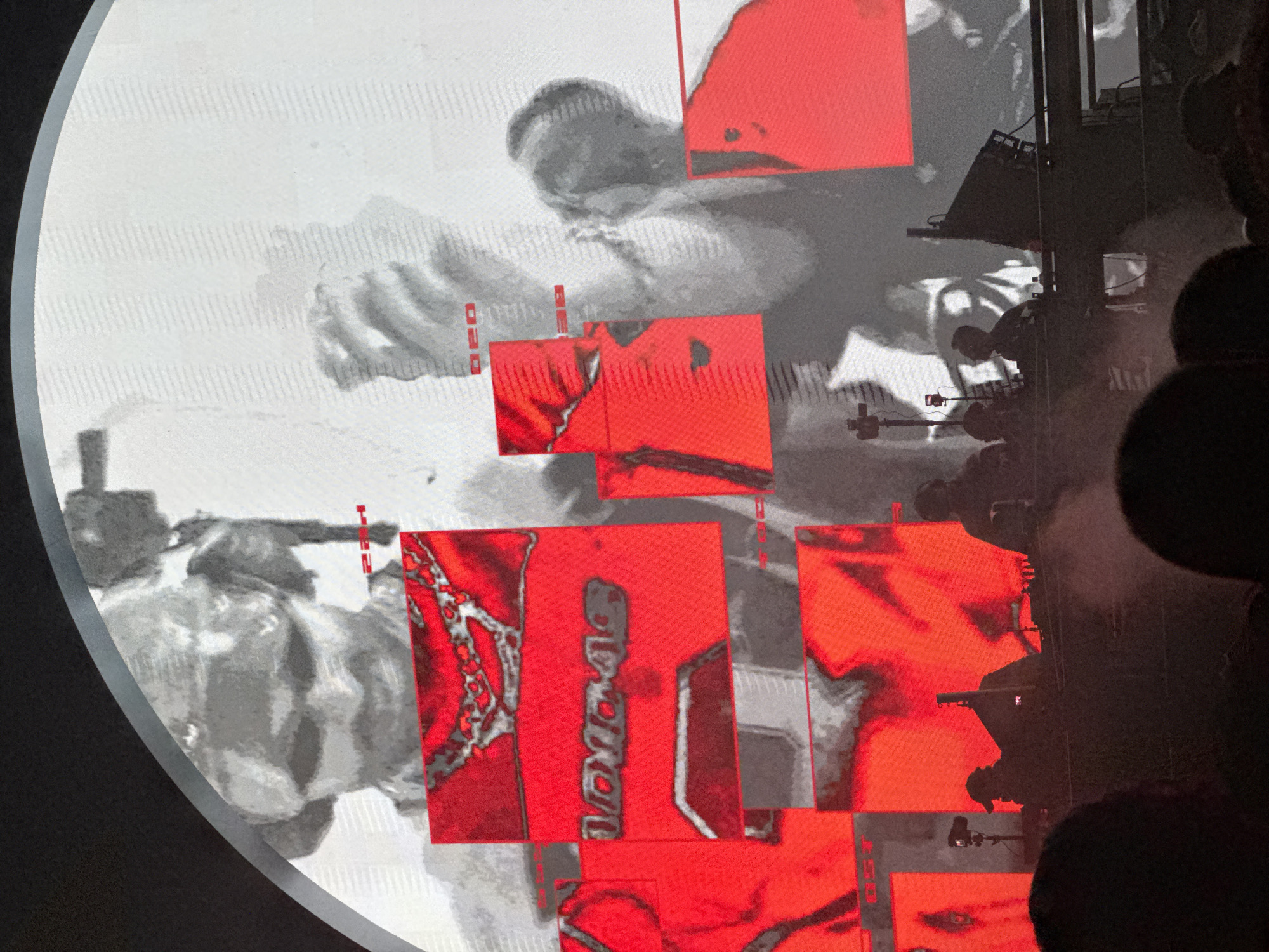 The image depicts a concert or event with a large screen displaying abstract red graphics and a black and white image, possibly a person. In the foreground, silhouettes of people are visible, some appearing to be DJs or performers at a console. The overall atmosphere suggests a vibrant, live performance. The event took place in Sofia, Bulgaria.