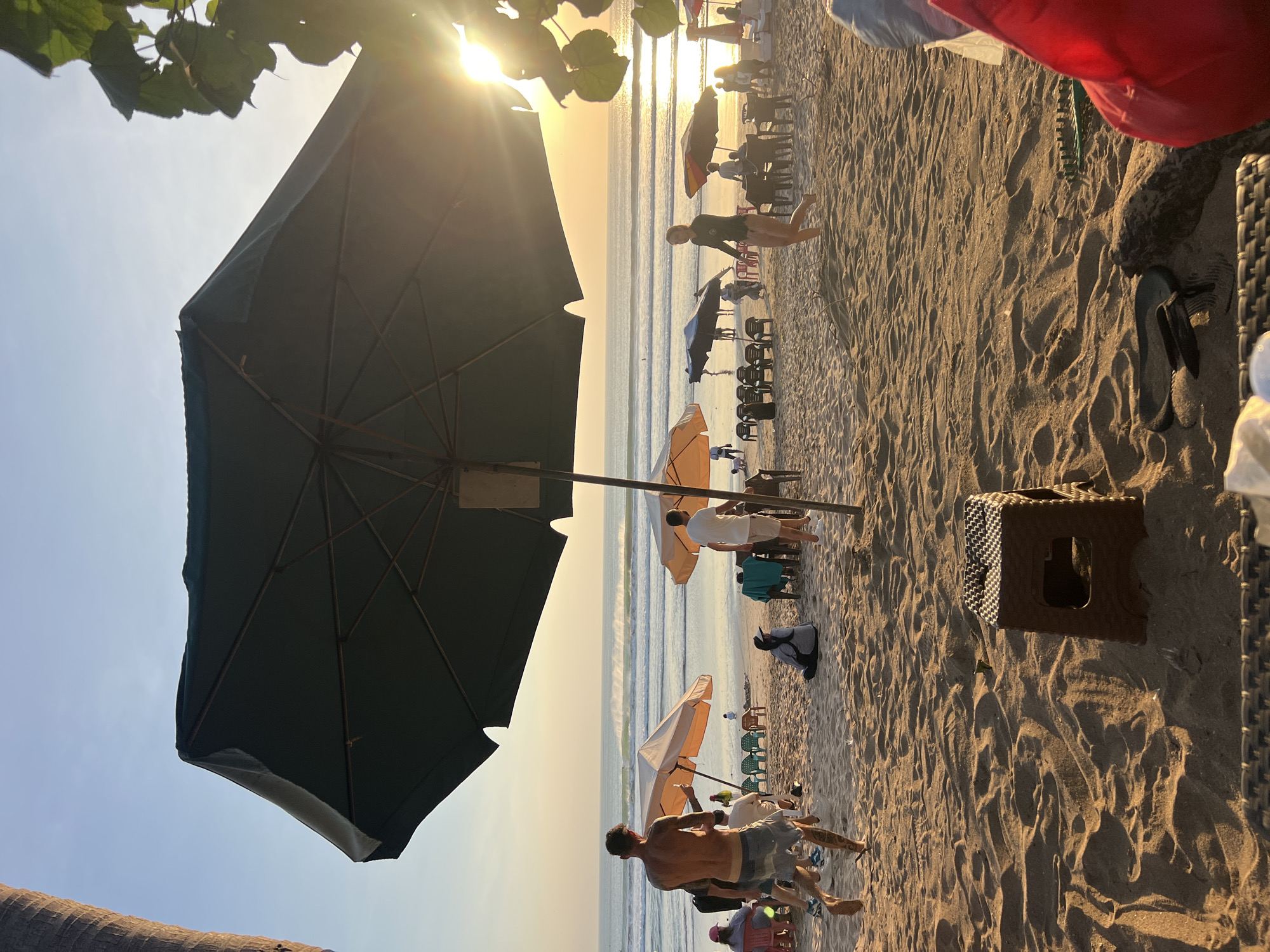 In Kuta, Indonesia, on a sandy beach at sunset, people are enjoying the evening. A large, dark green umbrella dominates the foreground, casting a shadow over the sand. In the mid-ground, several lighter-colored umbrellas are set up, with beach chairs and tables arranged around them. A few individuals are visible, some walking, others sitting or standing. The ocean is in the background, with gentle waves breaking on the shore. The setting sun casts a warm, golden light across the scene, creating a serene atmosphere.