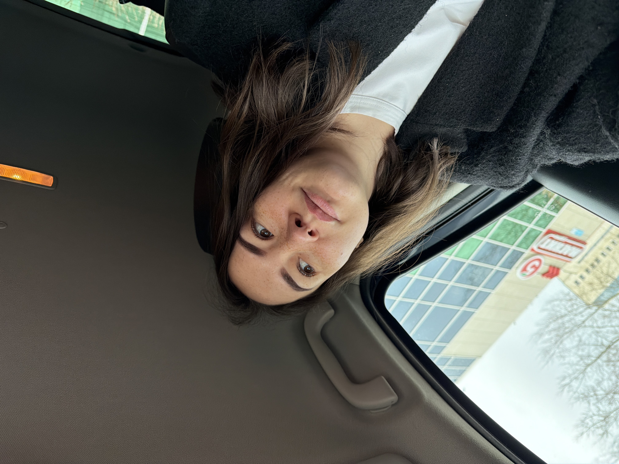 A woman is in the driver's seat of a car, looking at the camera. She has brown hair and freckles, wearing a white shirt and a black coat. The car's interior is visible, including the ceiling light, the car's ceiling, and a passenger assist handle. Outside the car window, a multi-story building with a sign is seen; the sign appears to say "Пятерочка" ("Pyaterochka"), indicating this may be in Russia. Given the reference to Chaikovsky, Russia, the location is likely there. The trees are bare suggesting winter. The building has glass panels. The scene is somewhat upside down from the perspective of the image, but the overall image captures the woman's selfie-style shot from within the car.