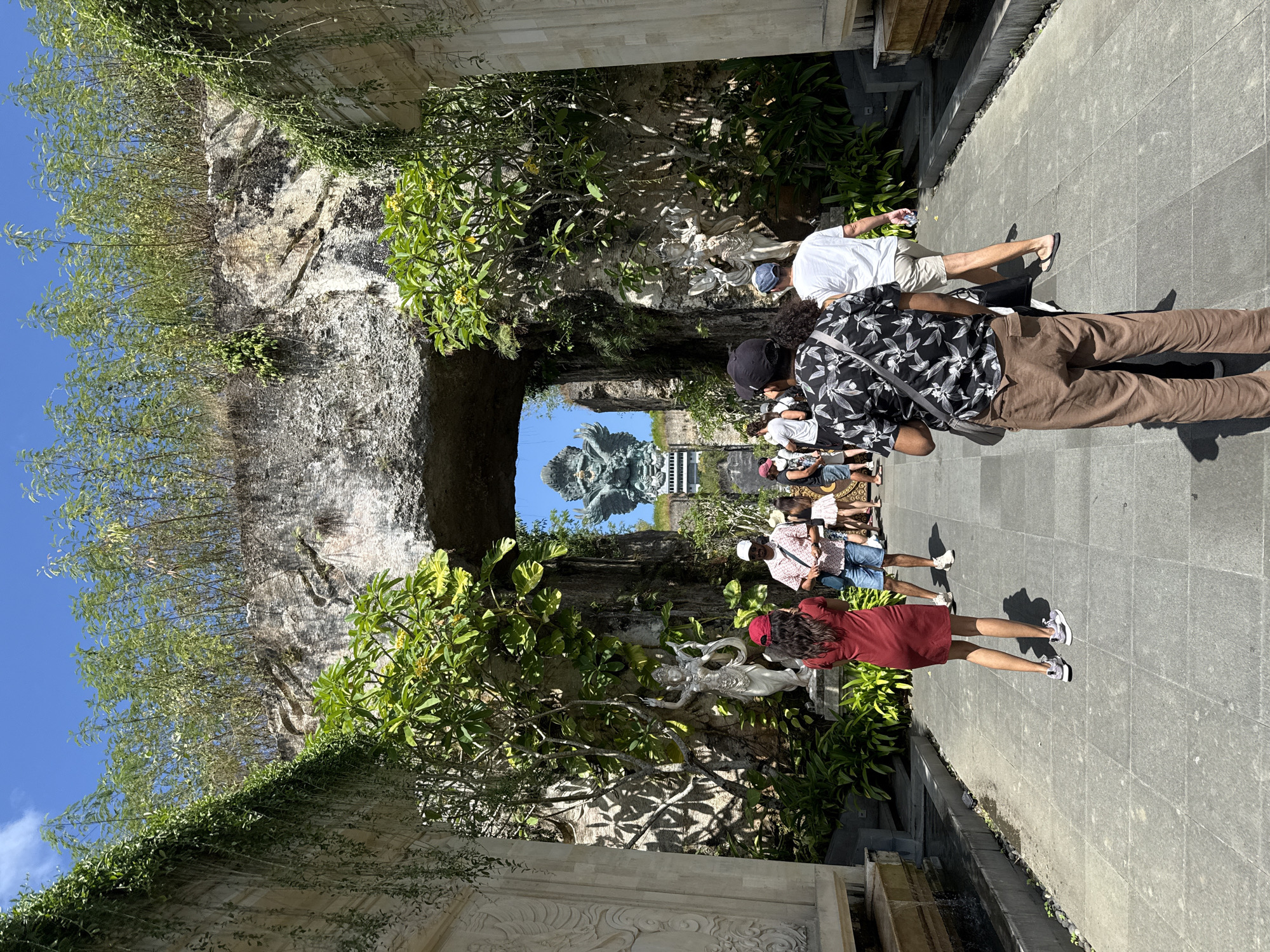 The image depicts a stone path with tourists walking towards a monumental statue in Kuta Selatan, Indonesia. The path is lined with carved stone walls, lush greenery, and statues. People are walking, some taking photos. In the distance, a large statue is visible against a clear blue sky.
