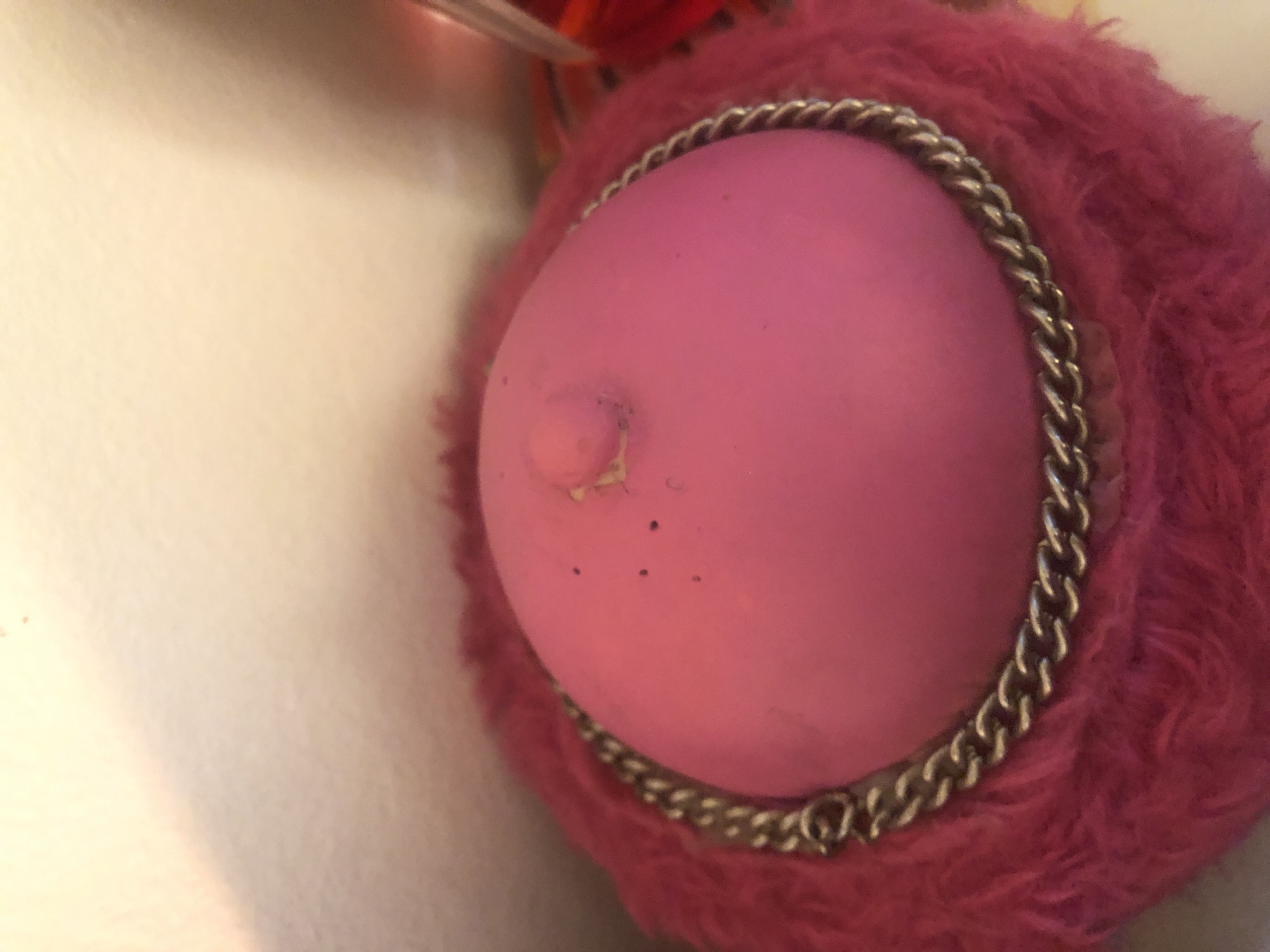 A close-up image shows a pink spherical object, possibly made of rubber or a similar material, surrounded by pink fur and a silver chain. The object has a raised nub on its surface and a few small dark spots. The background is a blurred off-white surface. The scene's location is not specified.