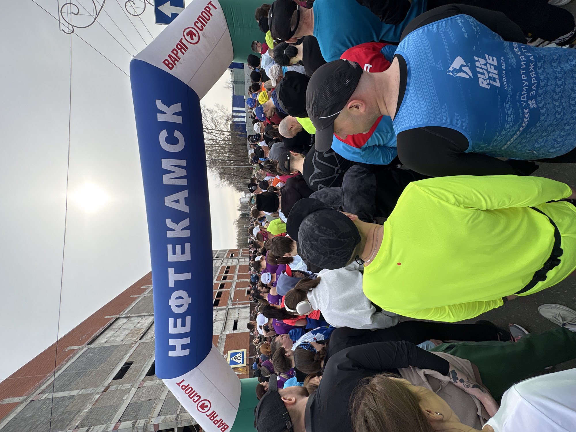 The image shows a large group of people gathered under an inflatable arch labeled "НЕФТЕКАМСК" in Cyrillic script, indicating the location is likely the city of Neftekamsk, Russia. The arch also bears the words "ВАРЯГ СПОРТ" in Cyrillic. The people appear to be participants in a running event, as one person is wearing a shirt with the English words "RUN LIFE" and "ШКОЛА БЕГА" (running school in Russian) printed on it. Another shirt reads "УДМУРТИЯ... ЗАРЯЖАЕТ" (Udmurtia Charges). The crowd is a mix of ages and genders, and many are wearing athletic clothing. The background includes a large building and a partly cloudy sky.