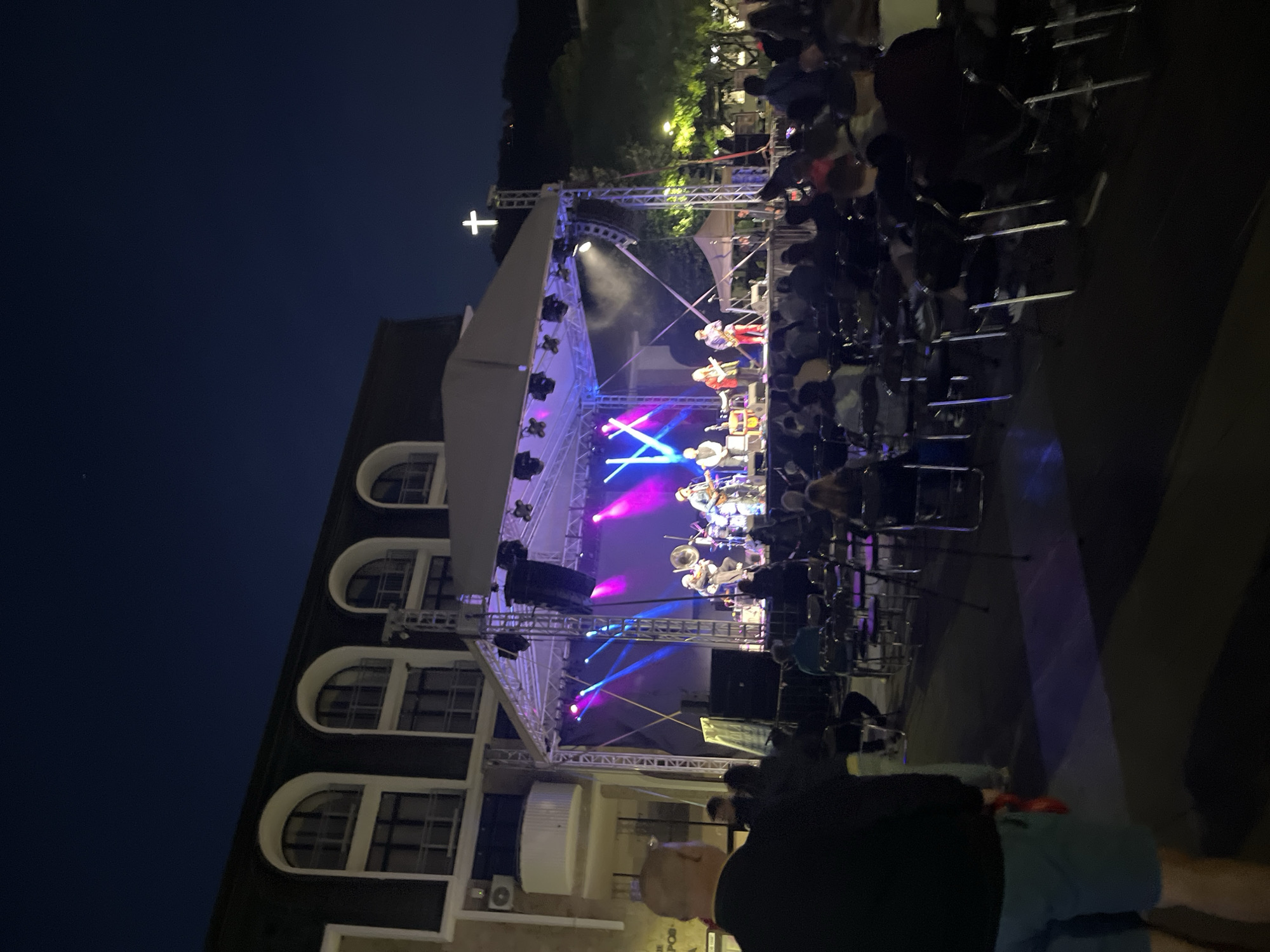 An outdoor concert is taking place at night in front of a building with arched windows. A band is performing on a stage with colorful lights. An audience is seated in chairs facing the stage. A cross is visible on a hill in the background.