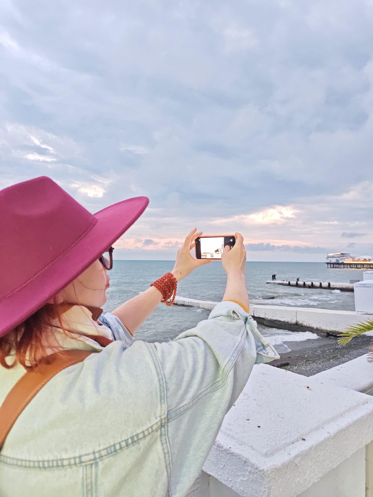 A woman with red hair, wearing a wide-brimmed pink hat, glasses, a light-colored denim jacket, and a beaded bracelet is holding an orange smartphone horizontally to take a picture of the sea and pier. She is standing on a white concrete wall. In the distance, the Sochi pier in Russia is visible with two figures standing on a wave breaker. The sea is calm with small waves, and the sky is cloudy with a hint of sunset. The image is taken from behind the woman, showing her focus on capturing the scene.