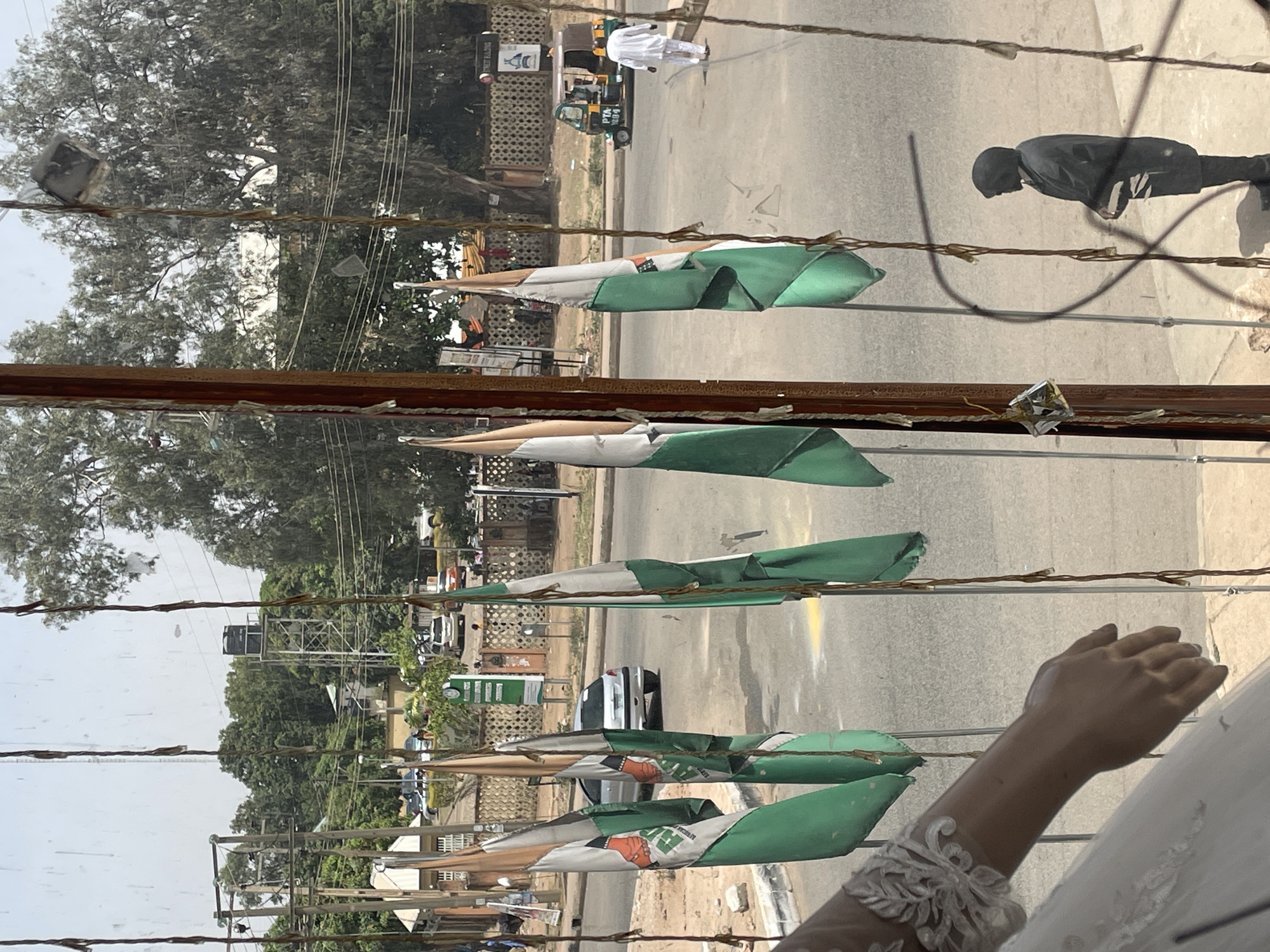 The image shows a street scene in Jos, Nigeria, viewed through a window. The street has several Nigerian flags, and some vehicles. People are walking on the side of the road. In the foreground, inside the building, there is a mannequin arm.